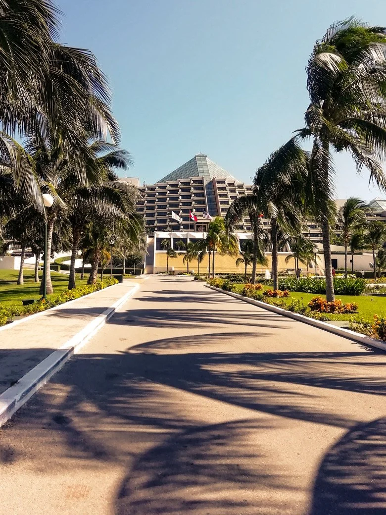 Un camino que lleva a un gran edificio con forma piramidal, rodeado de palmeras y jardines, con un cielo despejado.