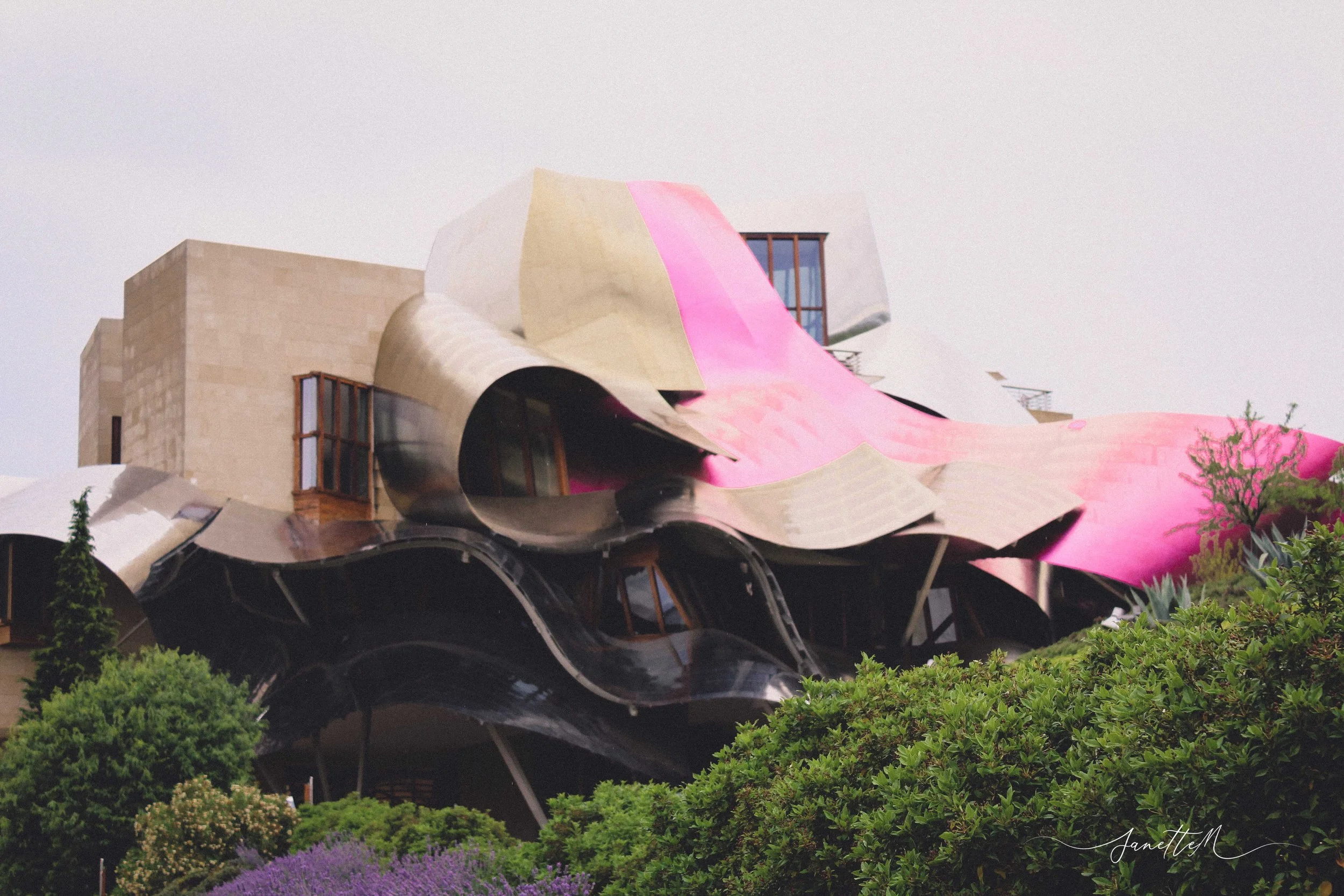 Edificio con estructura futurista y curvada, con colores metálicos y rosa, rodeado de vegetación y arbustos.