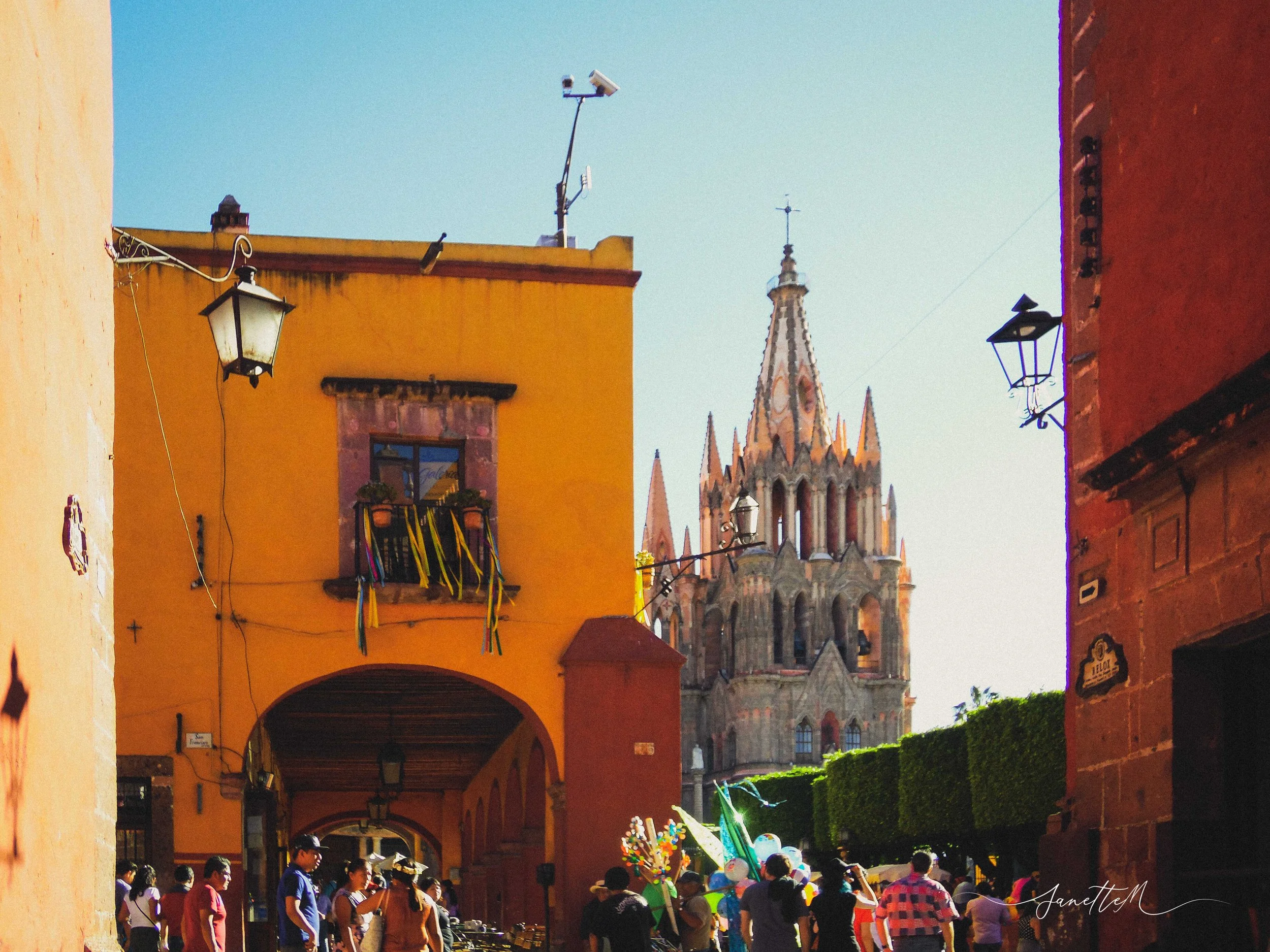 Calle colorida en un pueblo mexicano con personas caminando, vendedores ambulantes y una iglesia de estilo gótico en el fondo.