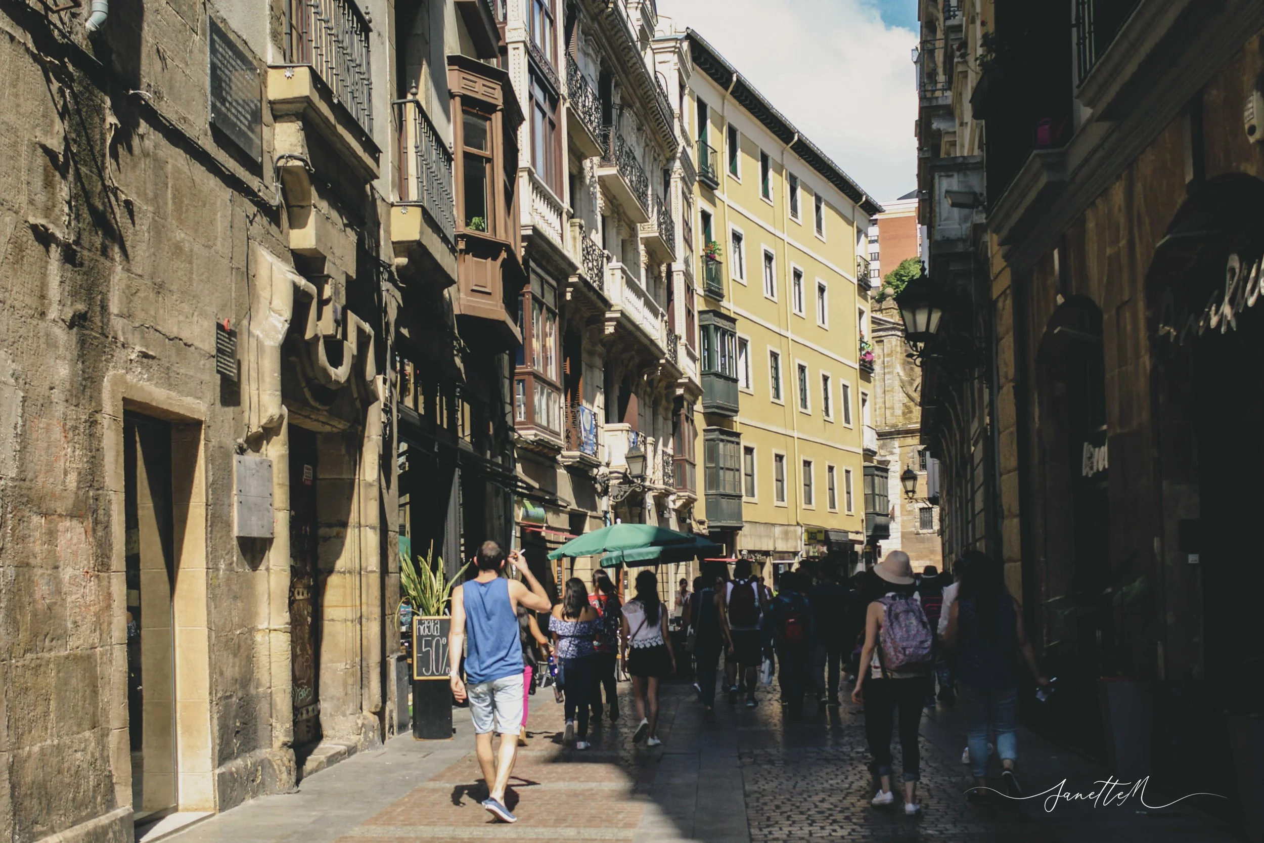 Persona caminando por una calle estrecha en una ciudad colonial con edificios de estilo europeo, algunos con balcones de hierro y fachadas coloridas, durante el día.