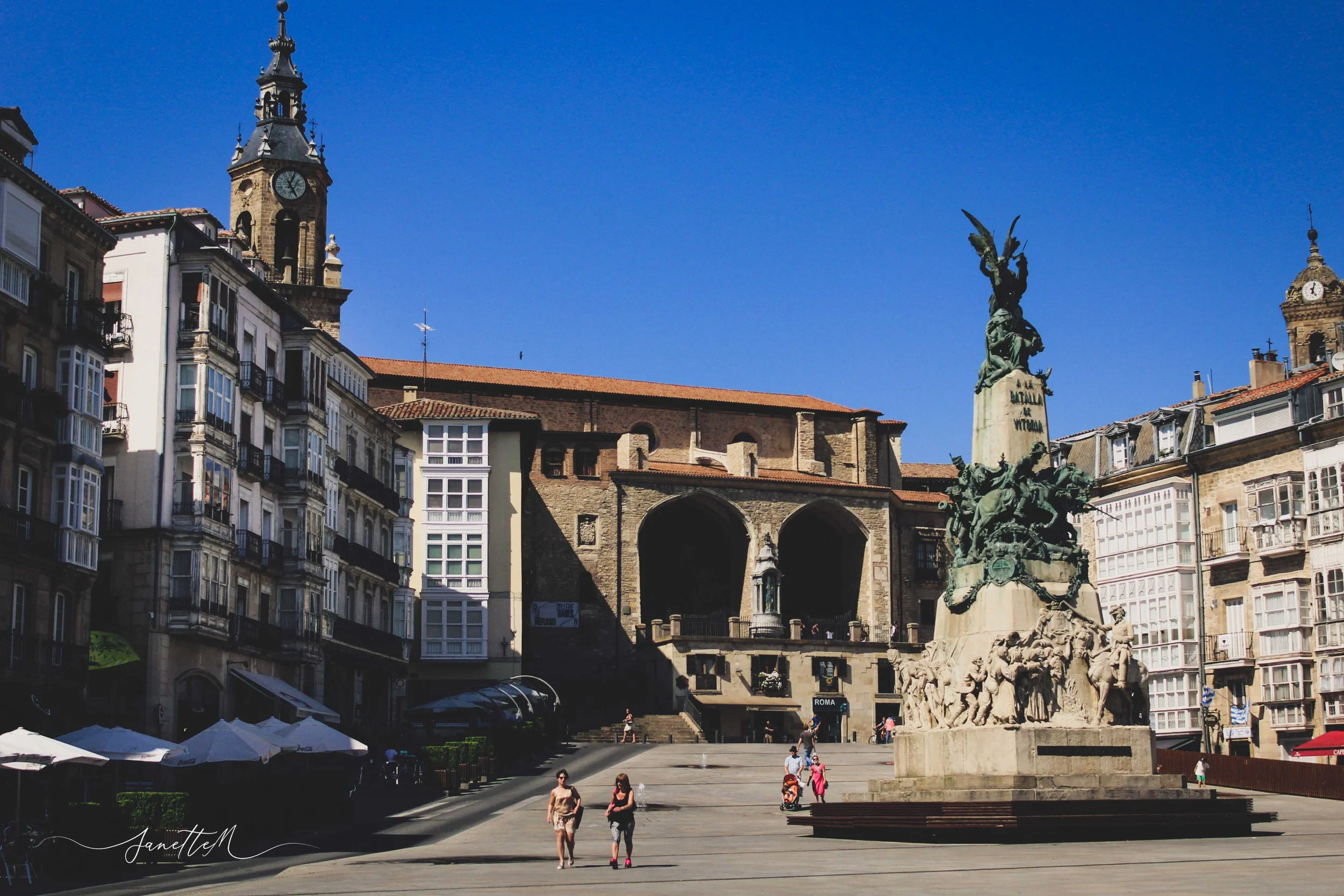 Plaza con una figura monumental en el centro, edificios con balcones y ventanas a su alrededor, un reloj en una torre y un cielo despejado.