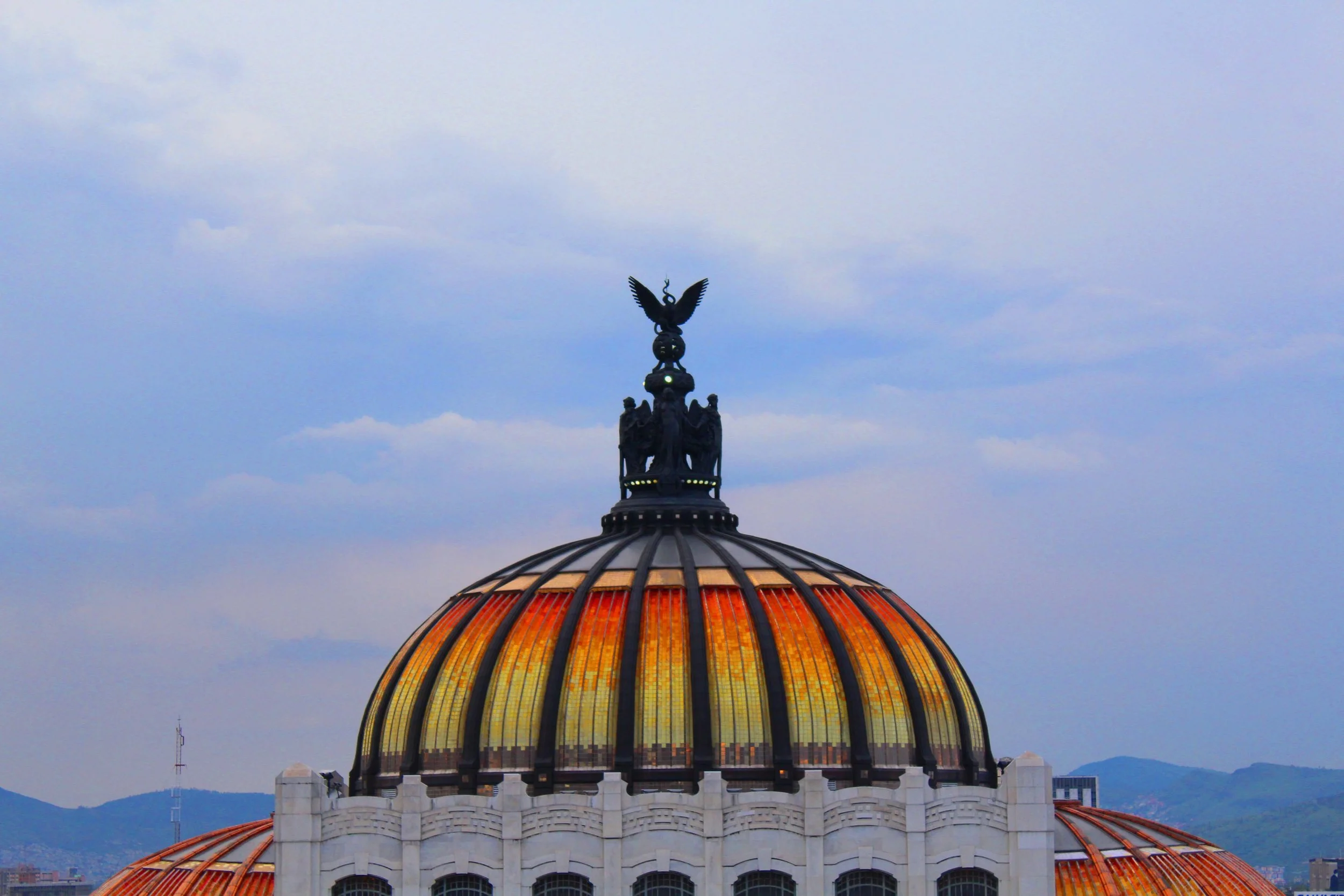 Cúpula de un edificio con ventana de cristales en tonos anaranjados y amarillos, con una escultura de un águila en la parte superior, contra un cielo con nubes.