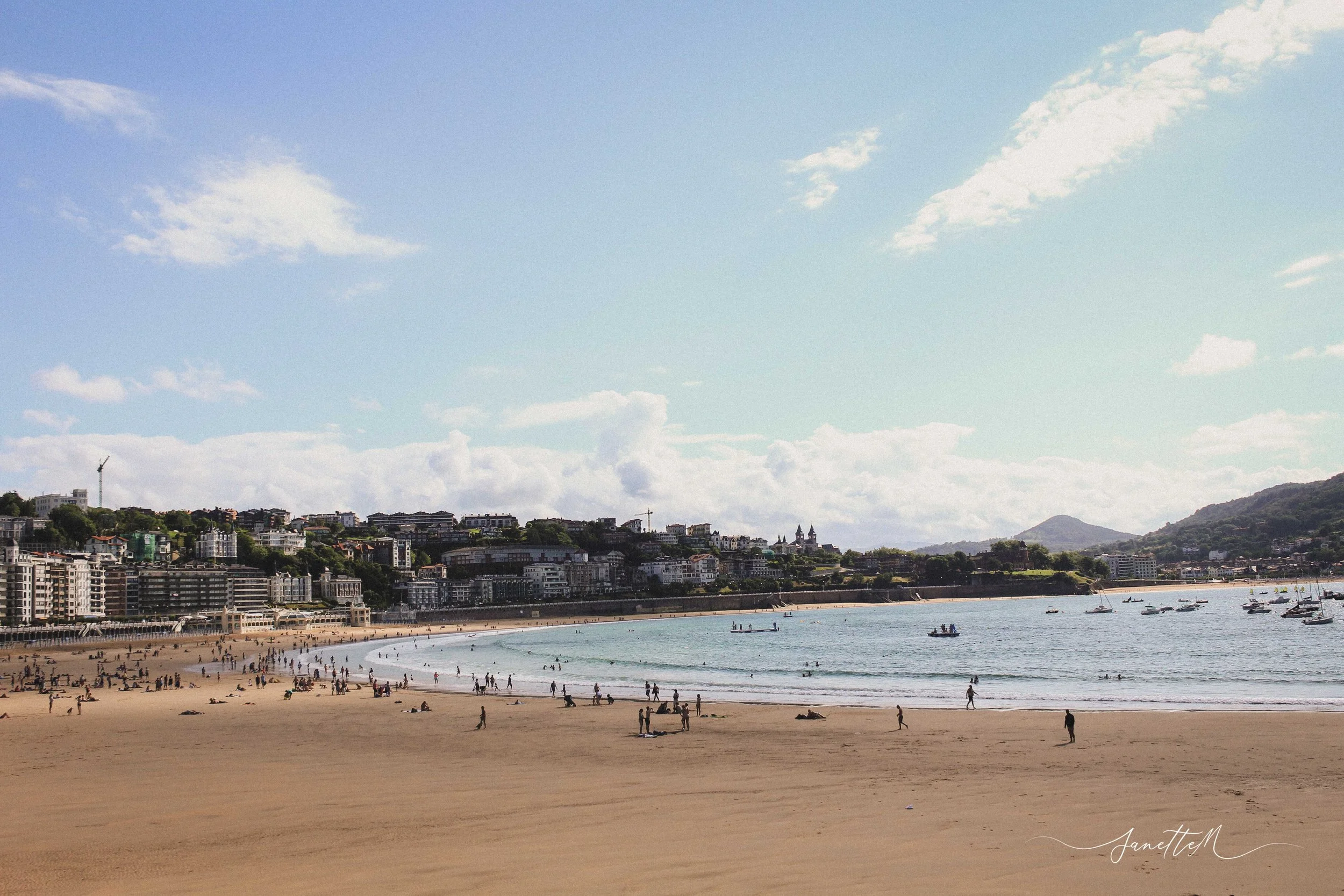 San Sebastián - Playa con arena dorada, personas caminando y disfrutando del mar, edificios y colinas al fondo, cielo azul con algunas nubes.