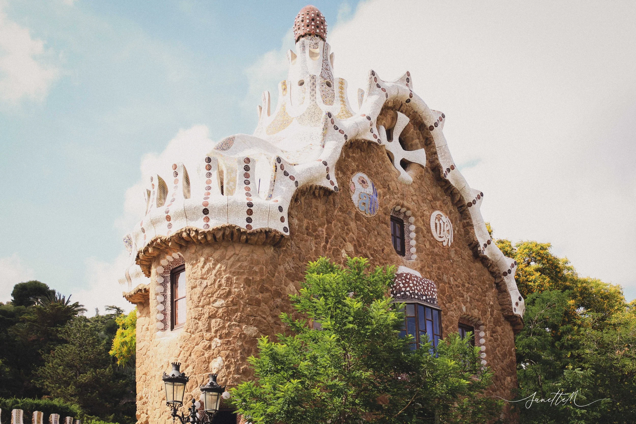 Barcelona - Parque Güell. Edificio de estilo modernista con fachada de piedra, paredes blancas onduladas y detalles de mosaico, rodeado de árboles y farolas, bajo un cielo con algunas nubes.