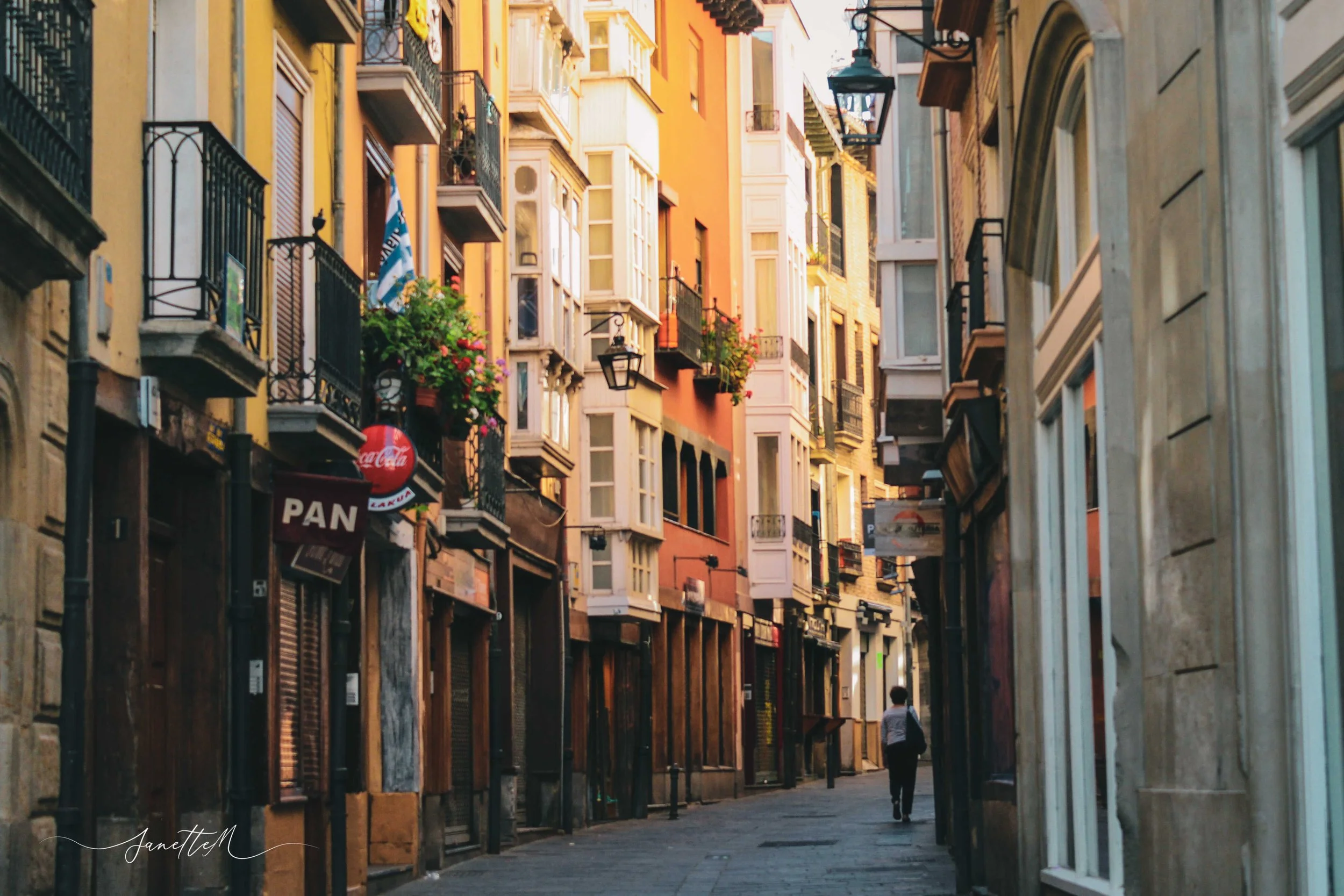 Una calle estrecha en una ciudad con edificios de colores cálidos, balcones con flores y toldos. Solo hay una persona caminando en la calle.