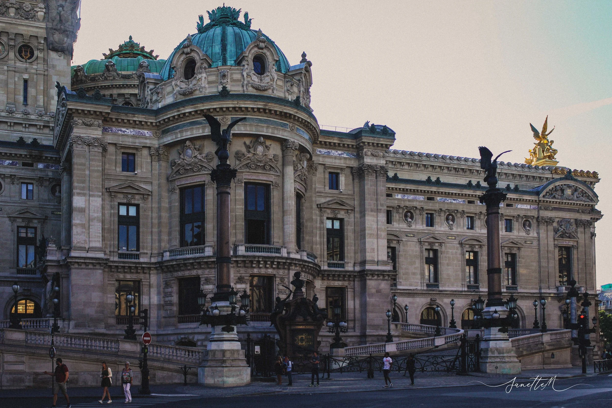 Edificio histórico con cúpulas azuladas y detalles ornamentales en estilo clásico. En la parte superior, hay estatuas doradas y columnas con esculturas de ángeles. En la calle frente al edificio, hay varias personas caminando y farolas negras decorat