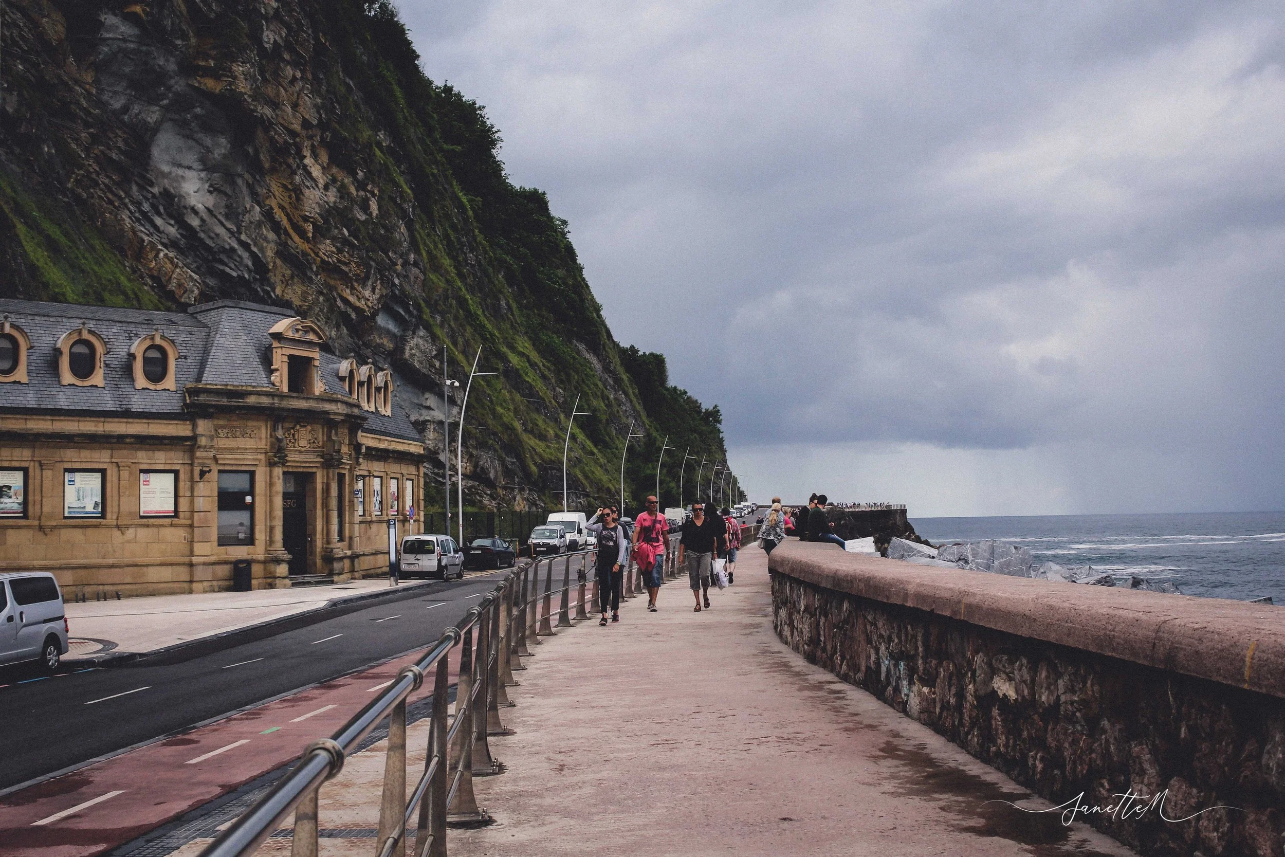San Sebastián - Camino peatonal junto al mar con personas caminando y un edificio antiguo a la izquierda, montañas a la izquierda y el océano a la derecha, en un día nublado.