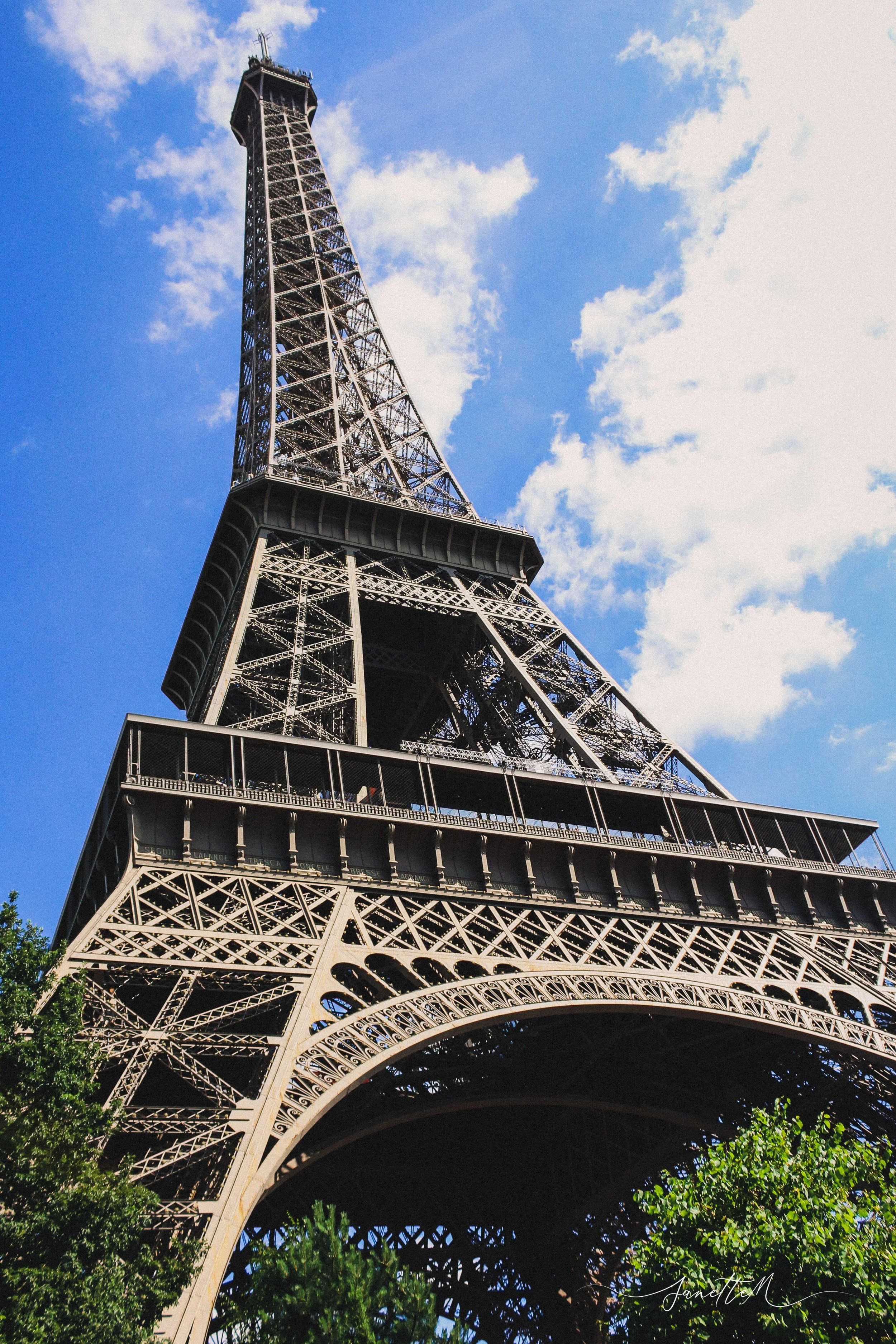 Vista en ángulo de la Torre Eiffel con cielo azul y nubes, incluyendo árboles verdes en primer plano.