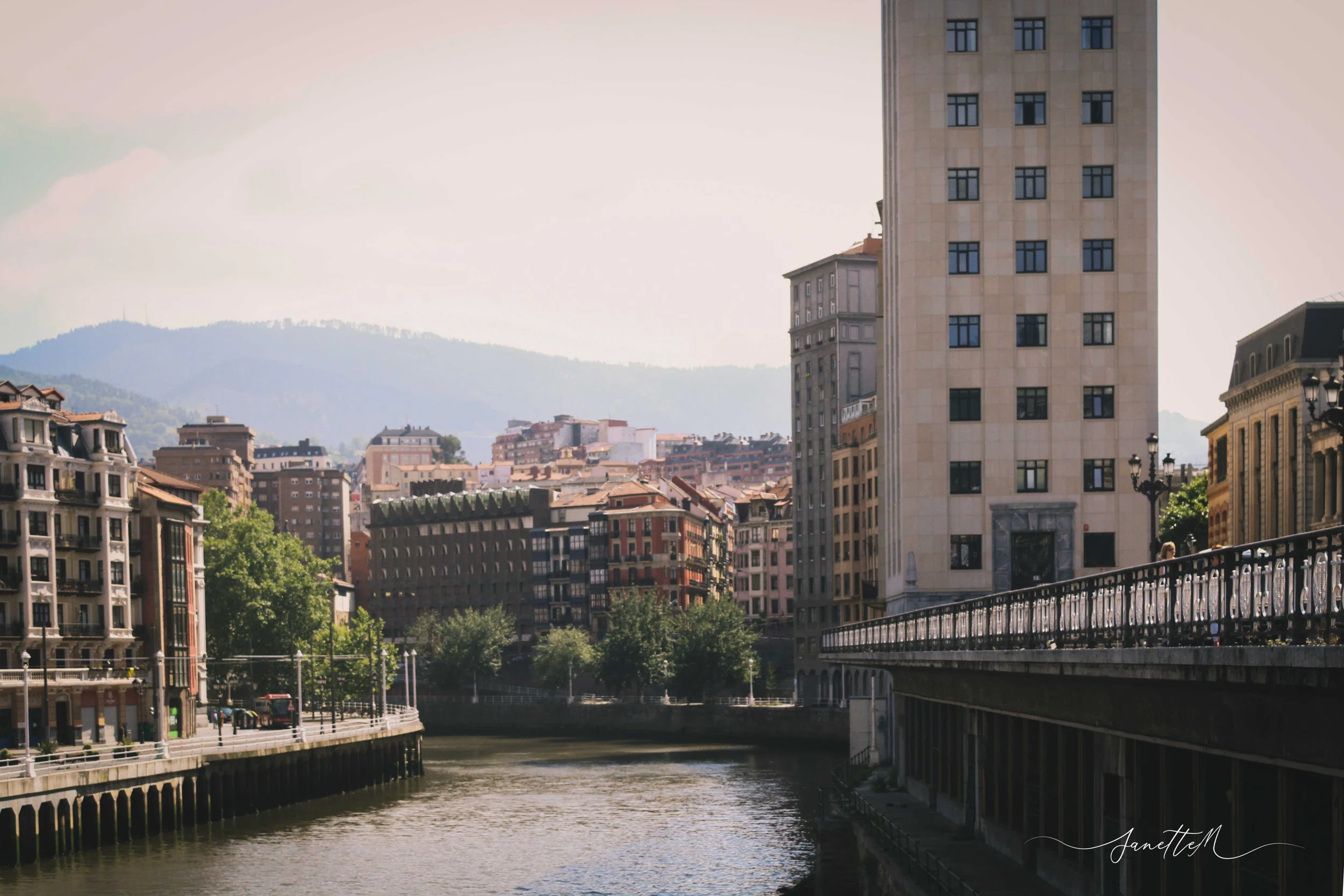 Vista de una ciudad junto a un río con edificios altos y montañas al fondo, día soleado.