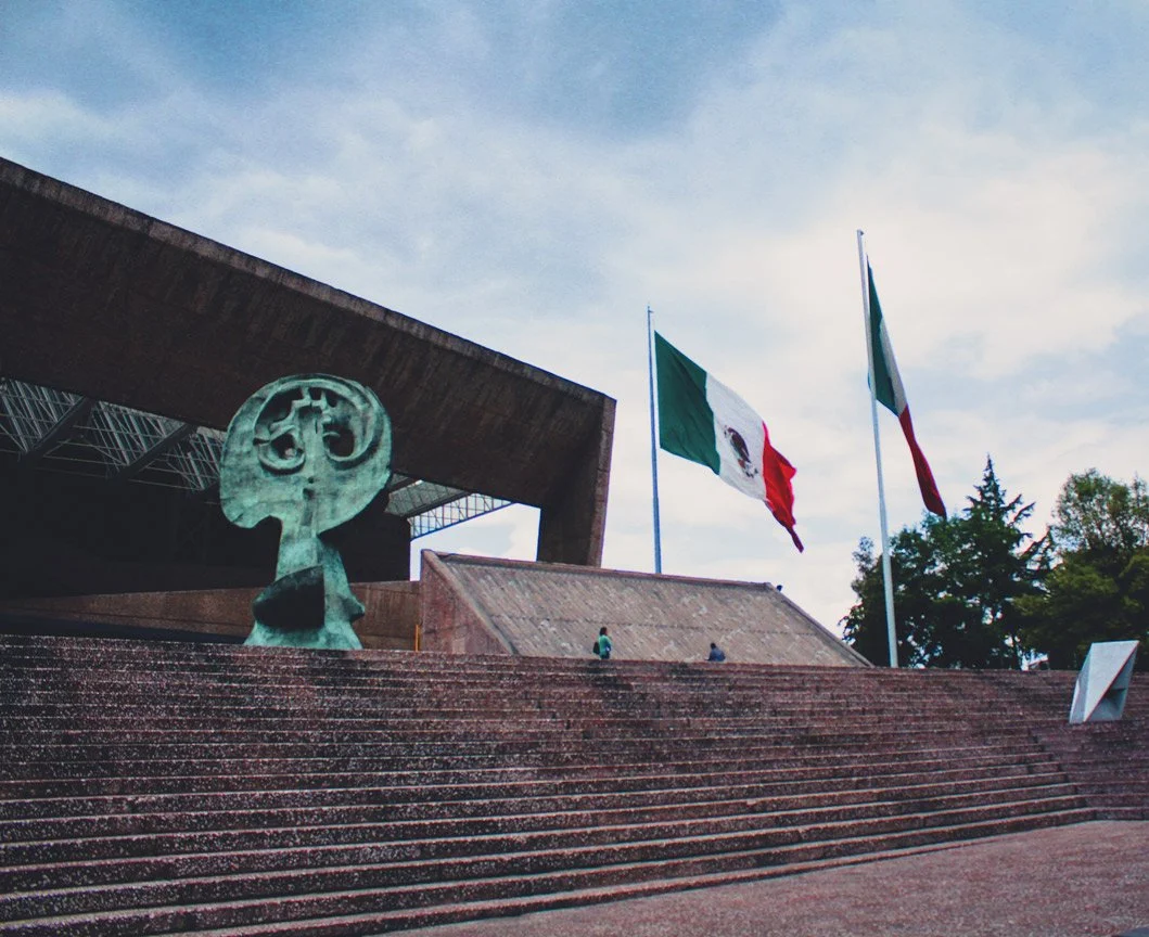 Escultura moderna en la entrada de un edificio, con banderas de México y una gran escultura de mal humor. Algunos visitantes suben las escaleras.