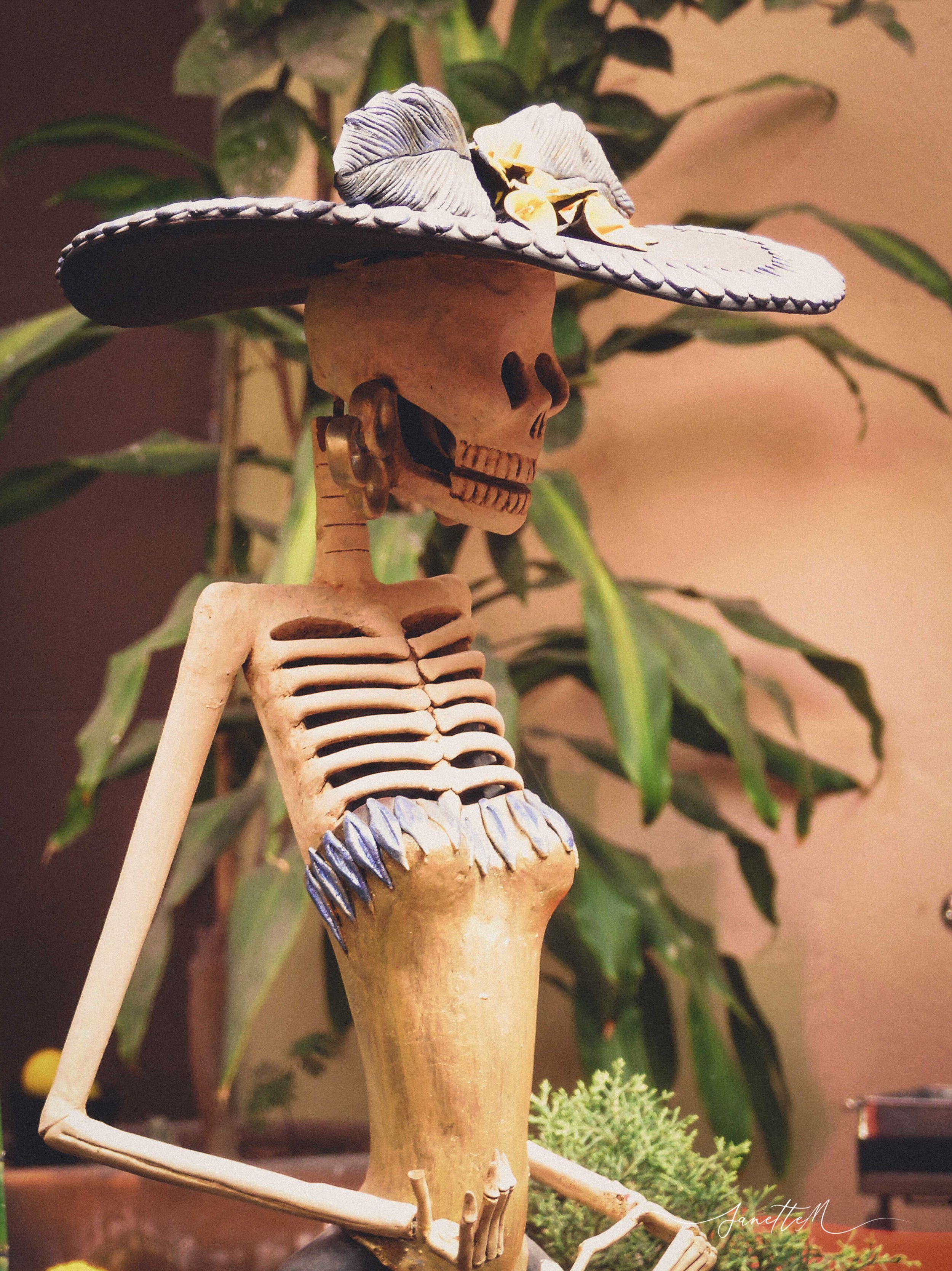 Escultura de un esqueleto con sombrero grande decorado con flores, rodeado de plantas verdes.