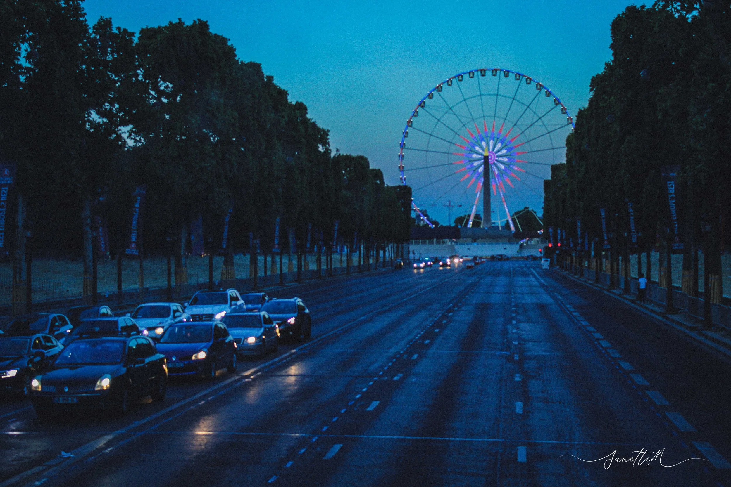 Vista de un parque de diversiones con una noria iluminada en la noche, al fondo, y un montón de autos estacionados y en movimiento en la calle frente al parque, rodeado de árboles.
