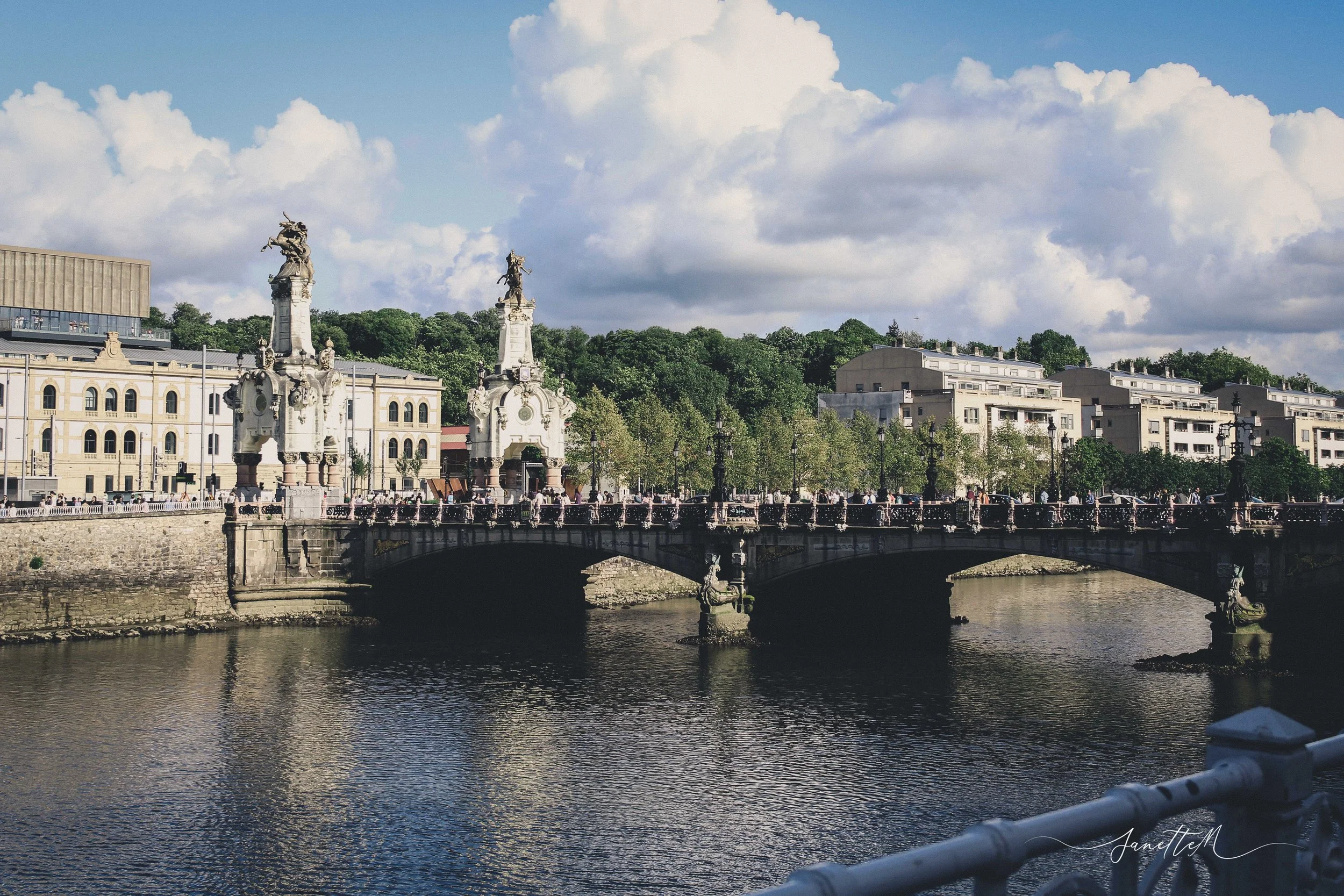San Sebastián - Puente con estatuas sobre un río, con edificios y árboles en el fondo y cielo nublado.