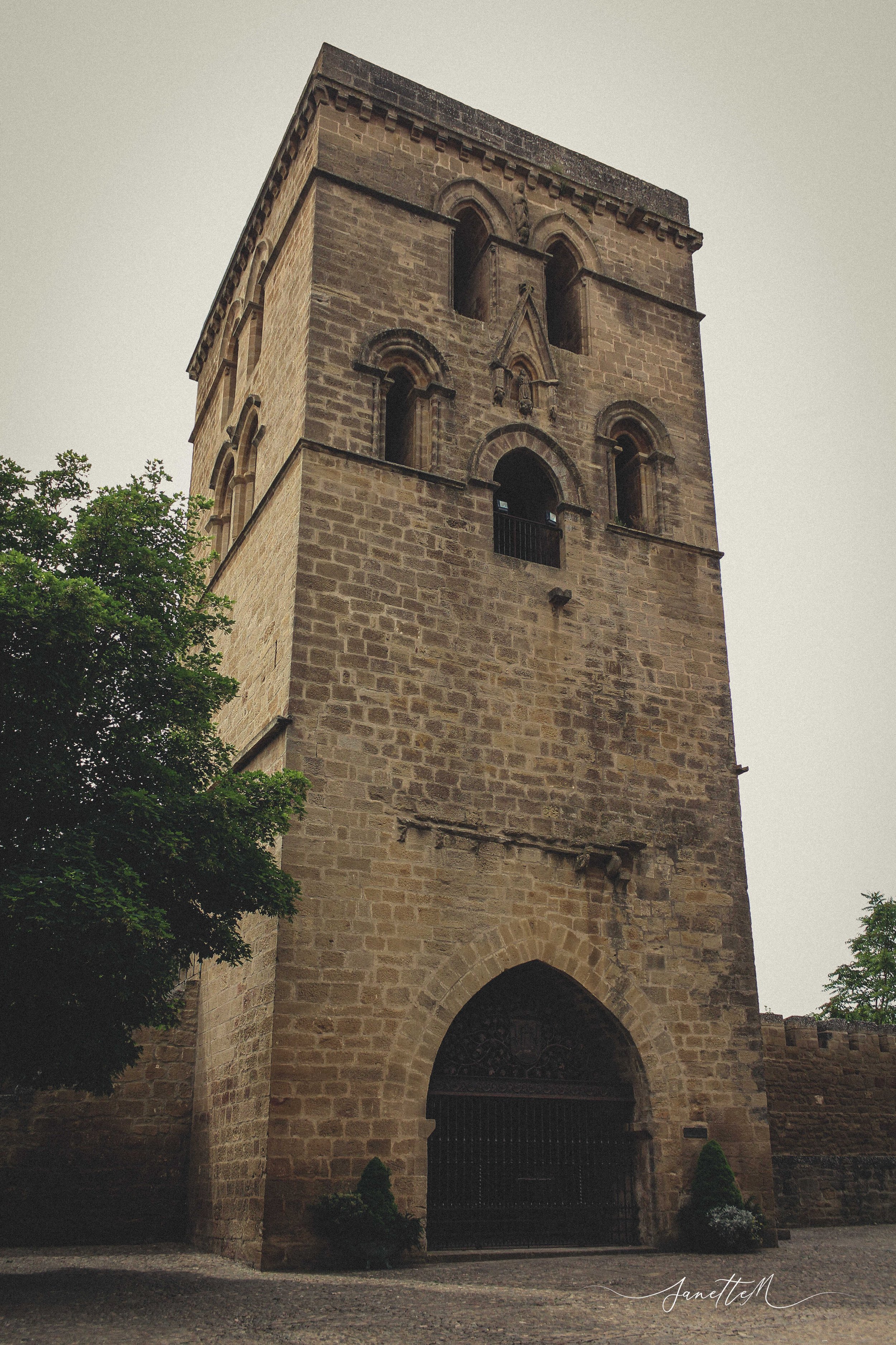 Torre antigua de piedra con ventanas arqueadas y entrada de arco en la base, rodeada de árboles y con un cielo nublado.