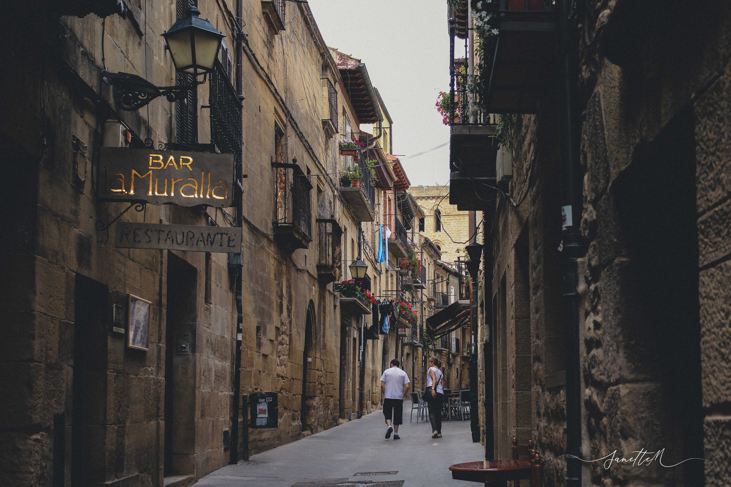 Vista de calle estrecha con edificios de piedra, balcones con flores y señal que dice 'Bar La Muralla' en una ciudad europea