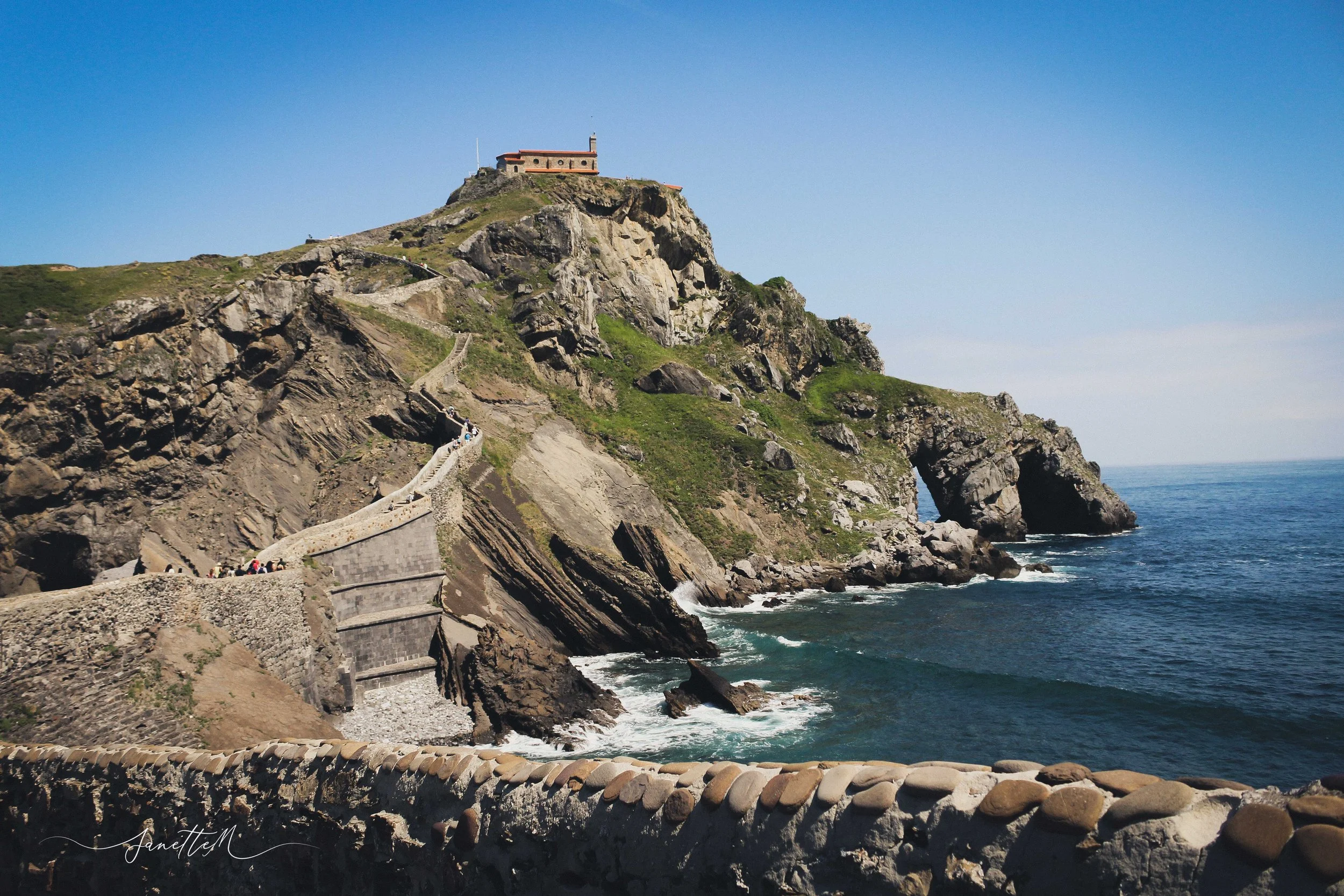 Vista del acantilado con camino y arteras en la costa, y una formación rocosa con un arco en el mar bajo un cielo despejado.
