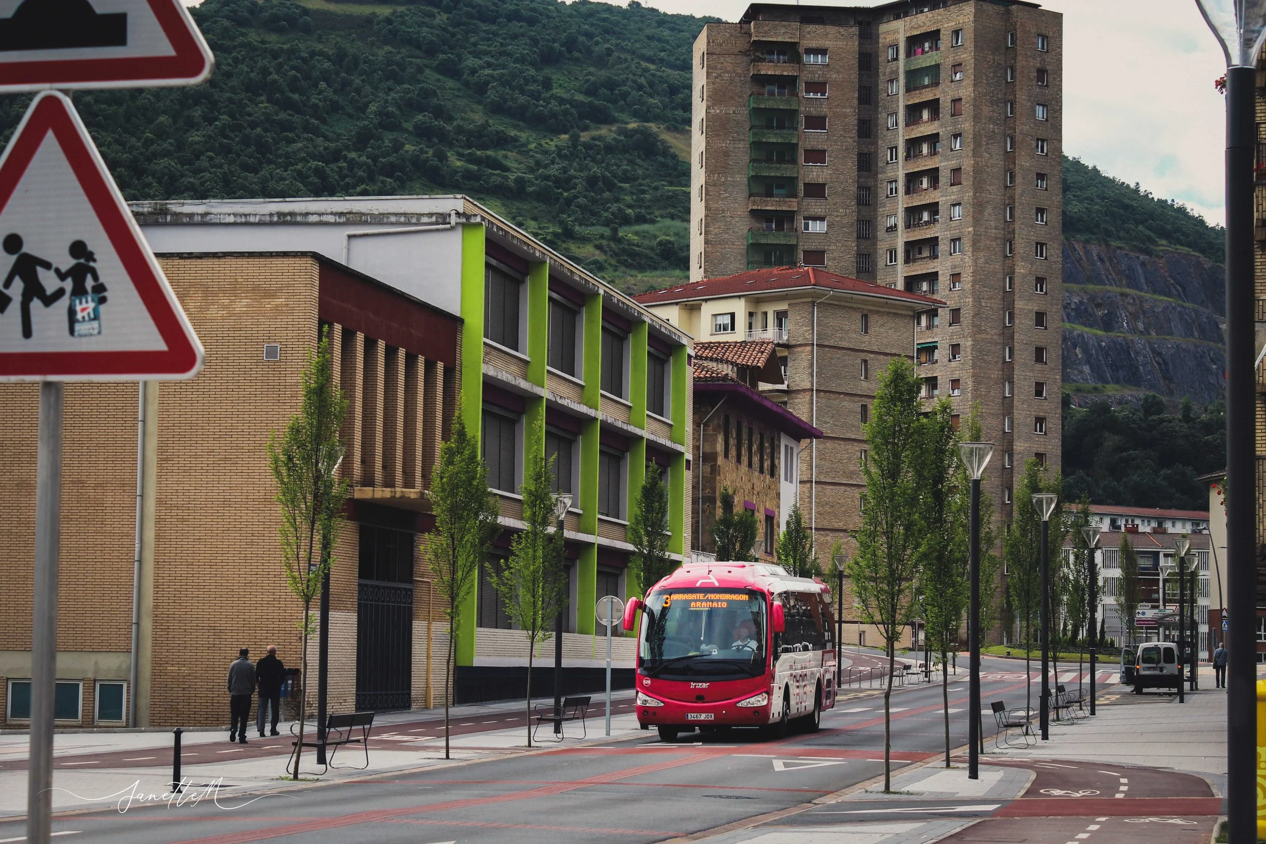 Vista de una calle urbanizada con árboles plantados y un autobús rojo, edificios residenciales y un fondo montañoso.