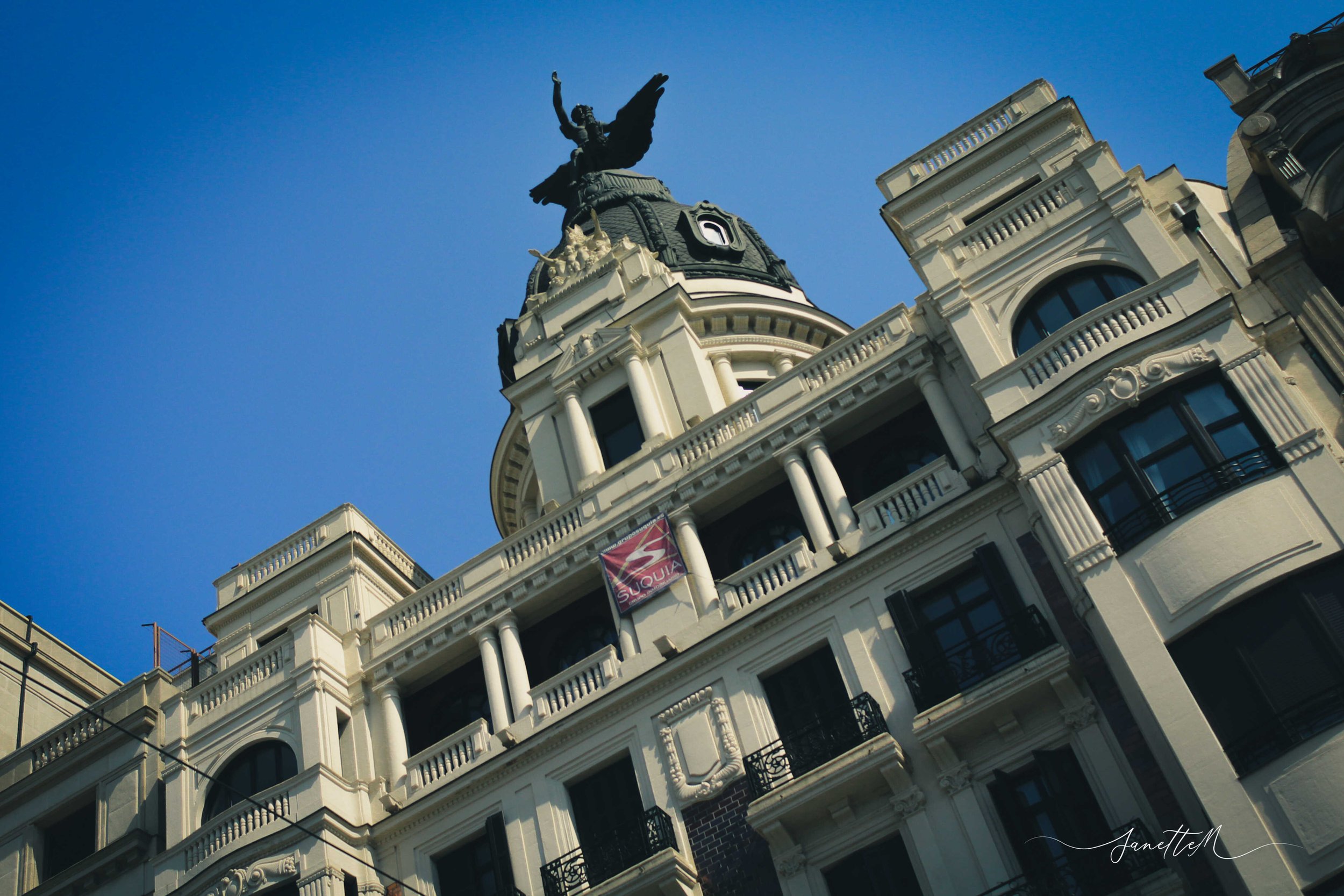Edificio antiguo blanco con muchas ventanas y balcones, con una cúpula y una estatua en la parte superior, contra un cielo azul.
