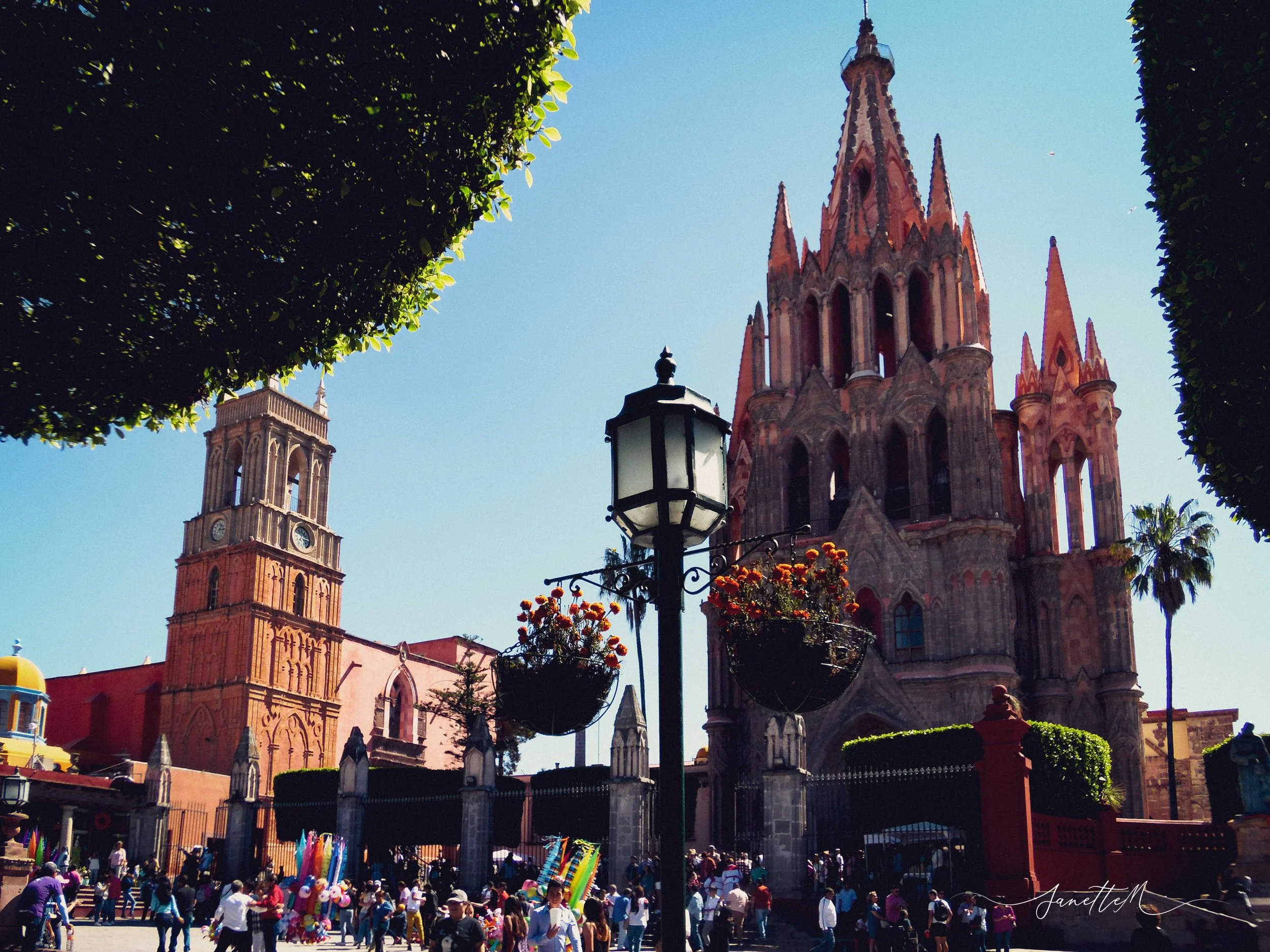 Plaza con personas, fuente, árboles y catedral gótica en el fondo, en un día soleado, con cielo despejado.