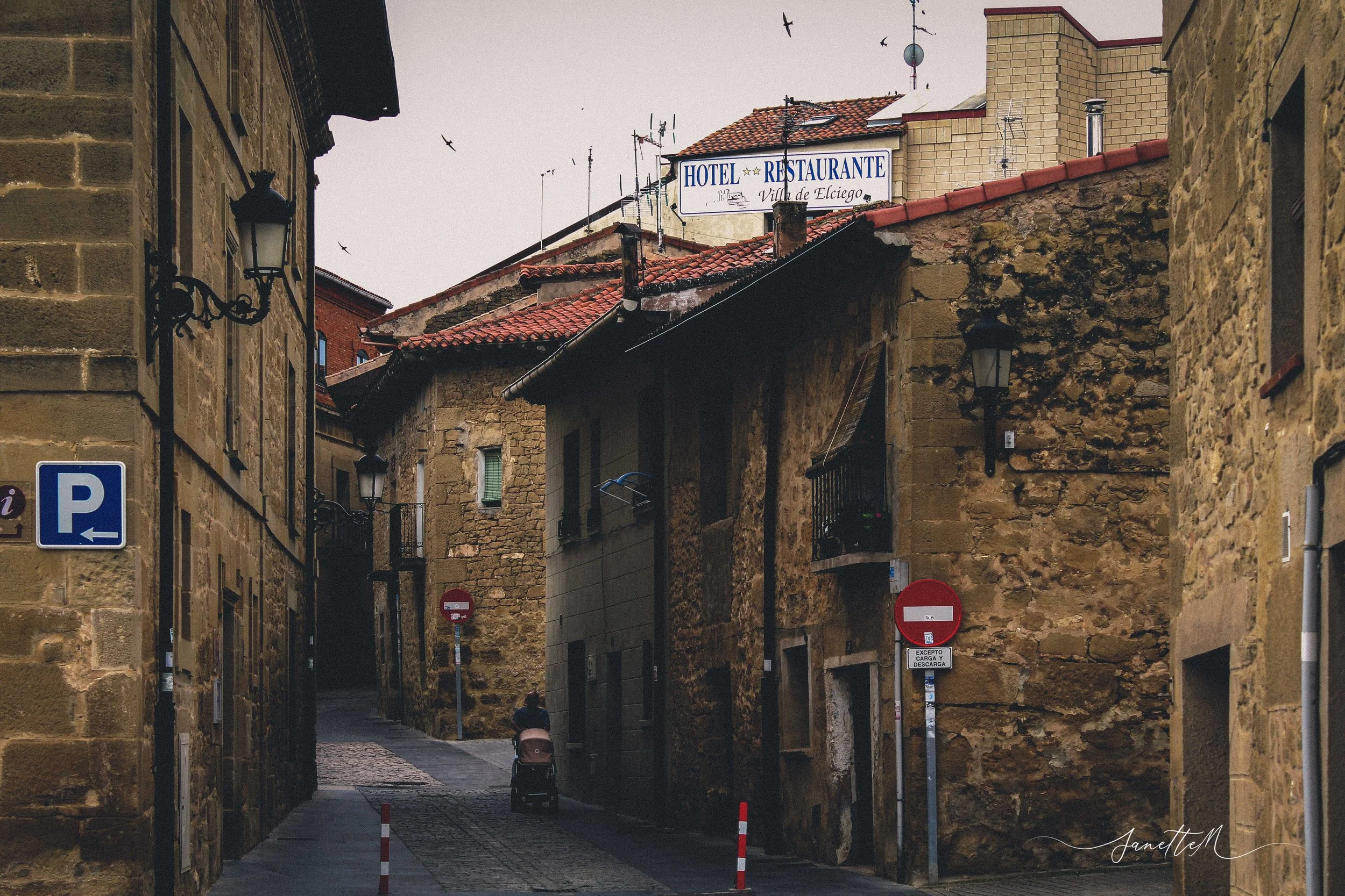 Calles estrechas con paredes de piedra, señal de 'P' para estacionamiento, señal de 'no entrar' y edificios con letreros de hotel y restaurante en un pueblo europeo.