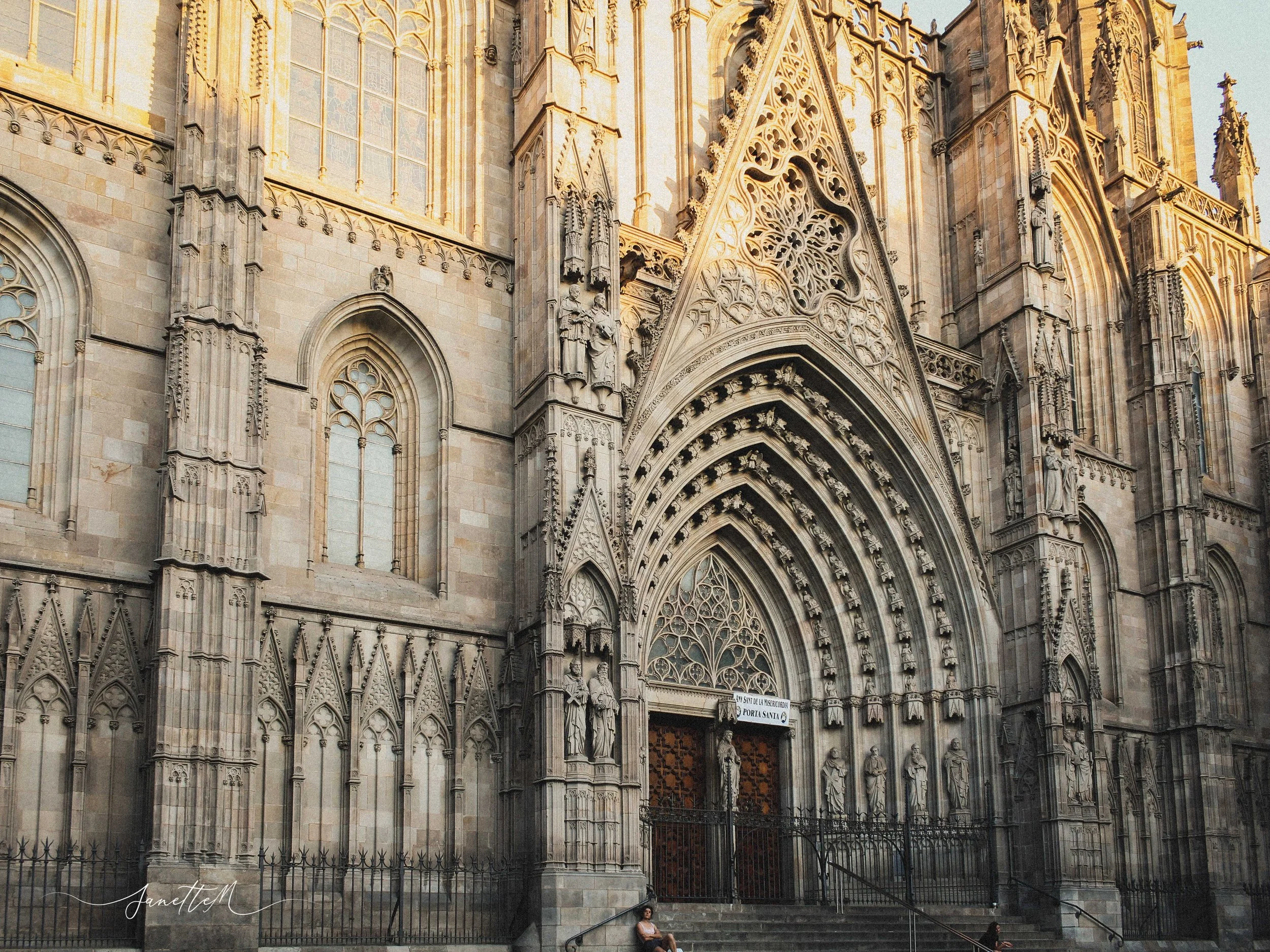 Barcelona - Fachada de una catedral gótica con detalles ornamentales y vitrales, entrada con arcos y estatuas sobre la puerta, al atardecer.