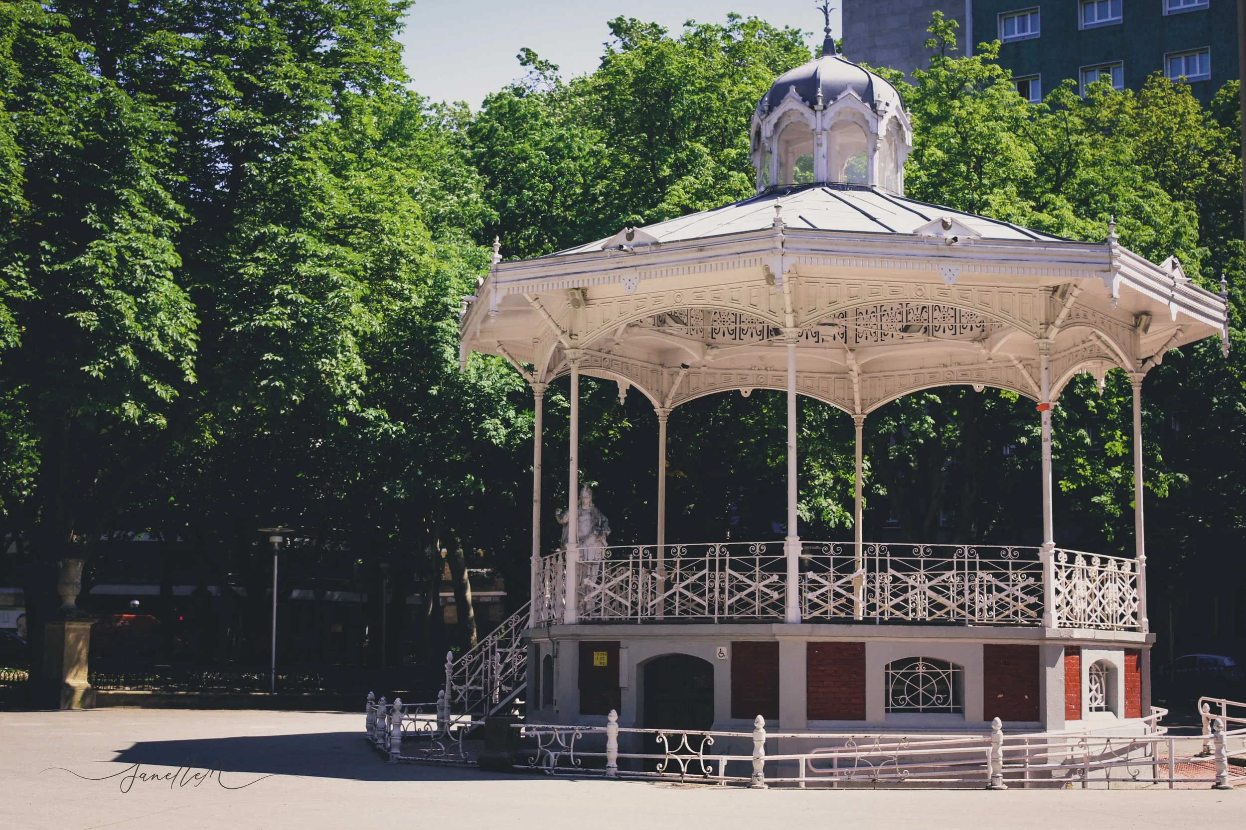 Quiosco en un parque con árboles verdes y edificios en el fondo, estructura blanca de hierro y escaleras.