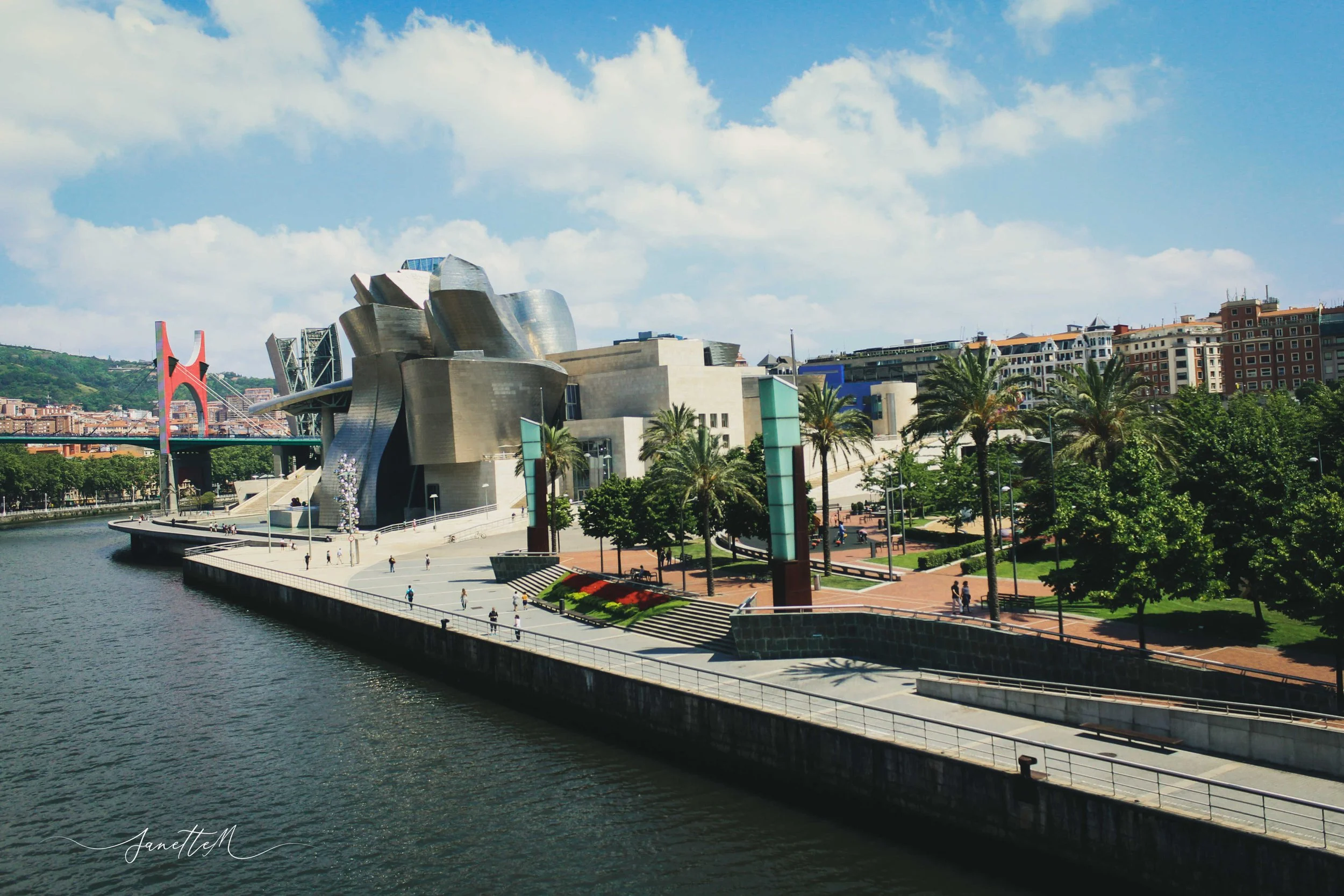 Vista del Museo Guggenheim en Bilbao, con río y parques en primer plano, y ciudad y puente en el fondo, en un día soleado con cielo parcialmente nublado.