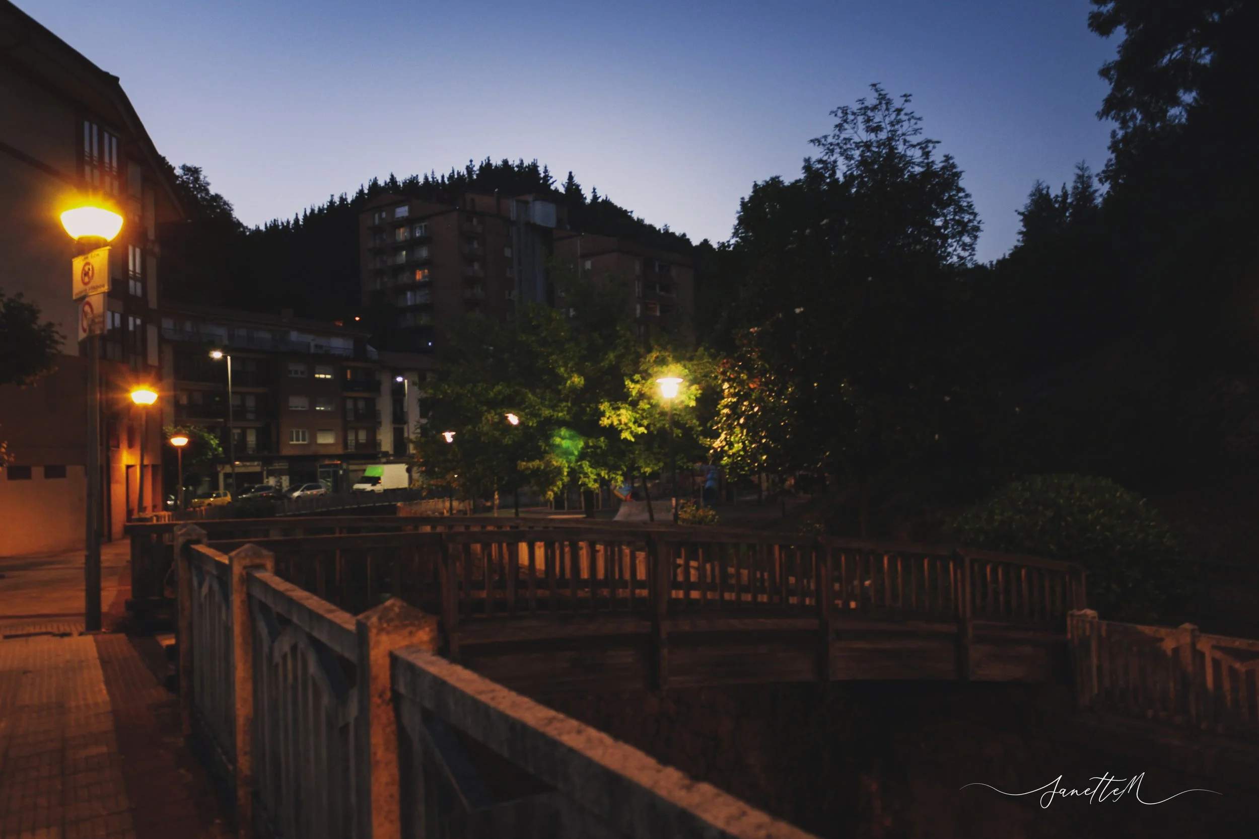 Puente de madera con barandales de madera en un parque urbano, iluminado por farolas, con edificios y árboles al atardecer o anochecer, en un entorno residencial