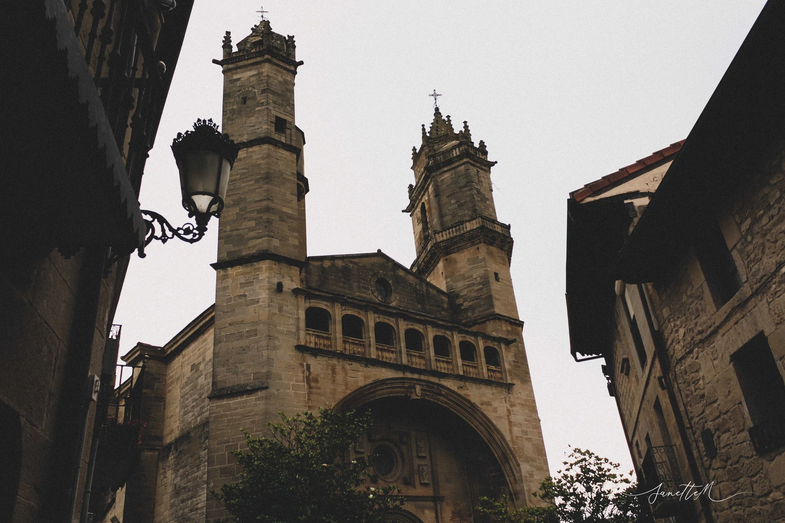 Vista en ángulo hacia una iglesia con dos torres en la ciudad, rodeada de edificios antiguos y árboles en primer plano, con cielo nublado.