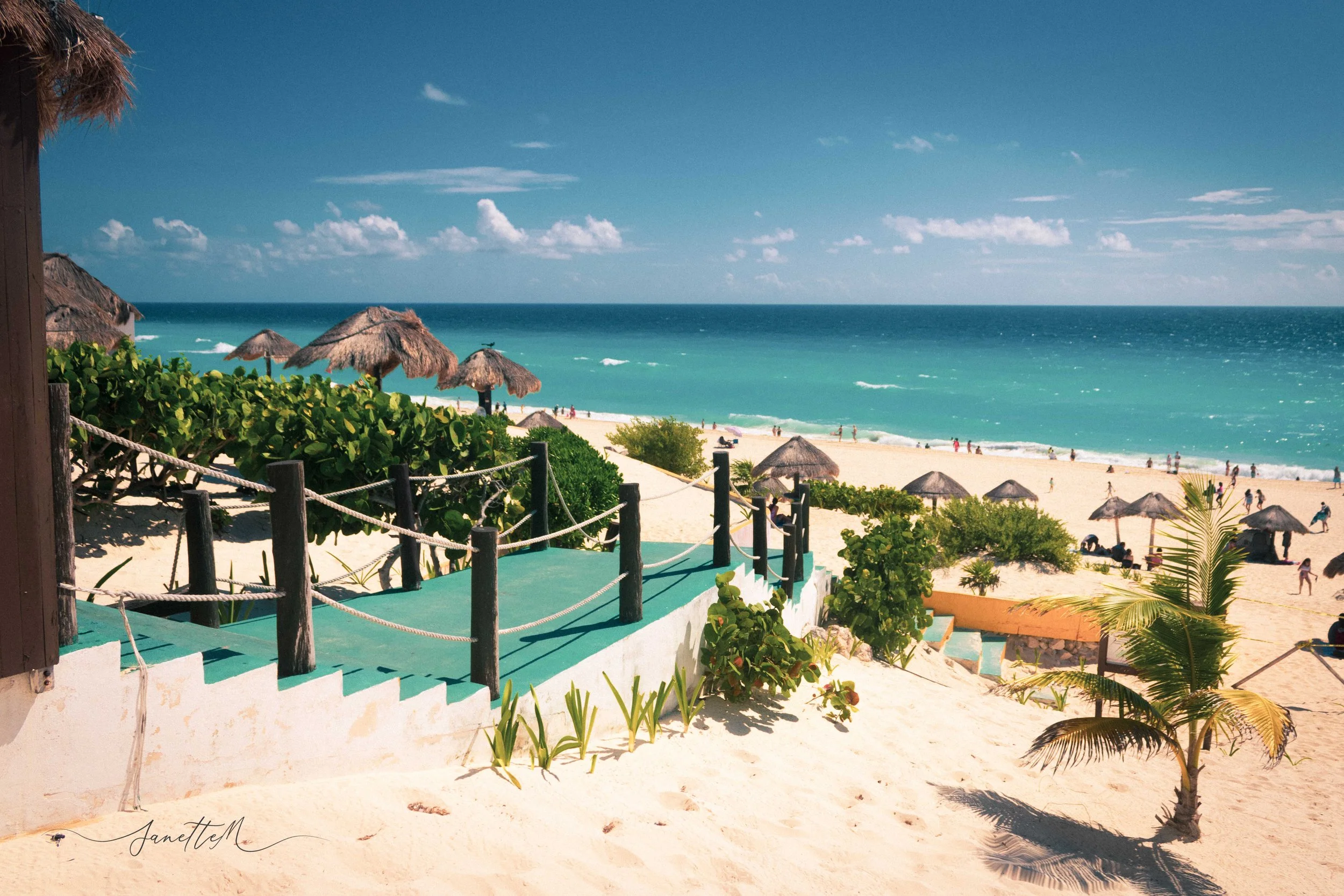 Vista de una playa con arena blanca, sombrillas de paja, vegetación y un camino elevado con barandilla de cuerda, frente al mar azul con olas pequeñas y cielo despejado con algunas nubes.