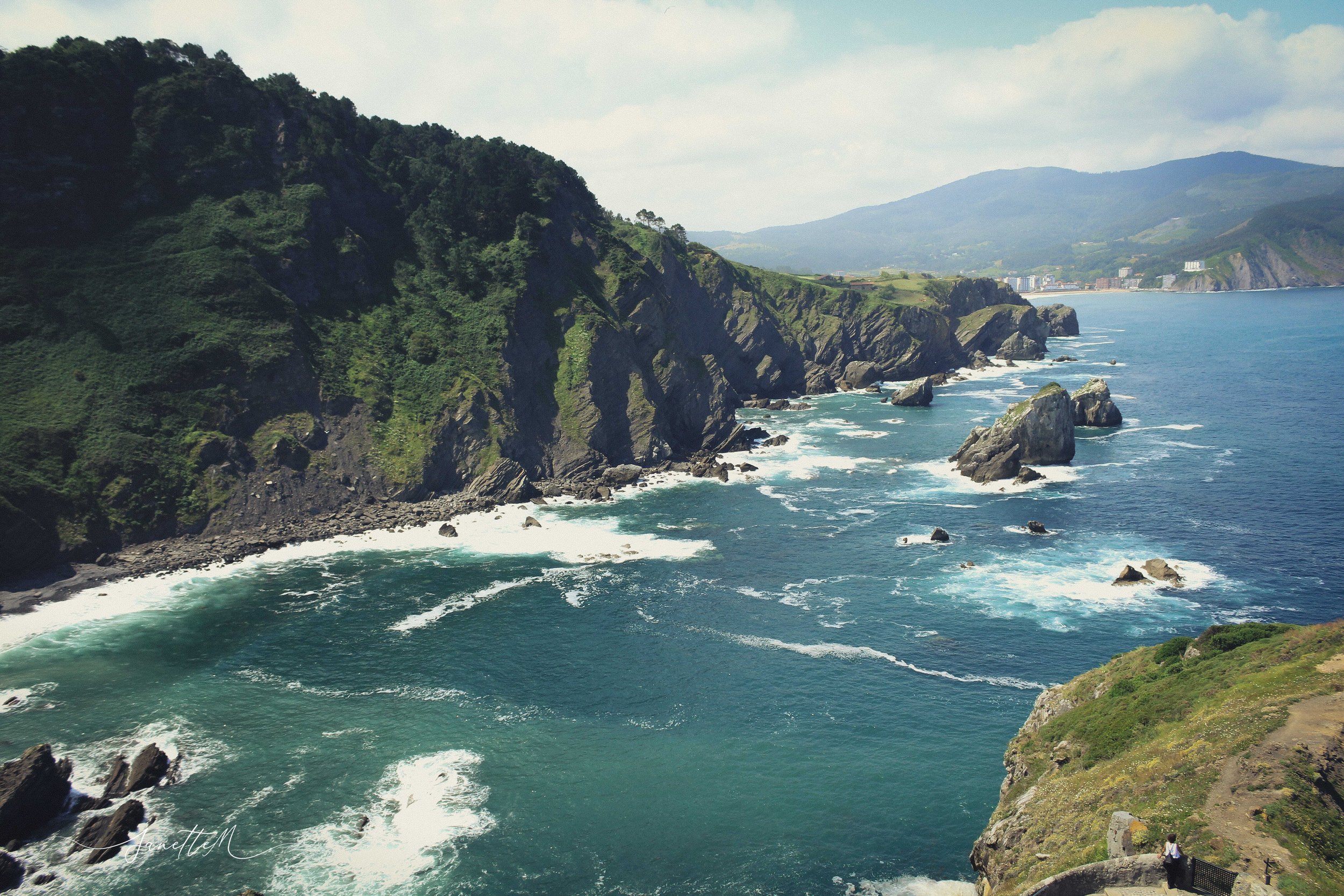 Costa escénica con acantilados verdes y rocas en el océano, vista desde un mirador con una persona en la esquina inferior derecha.