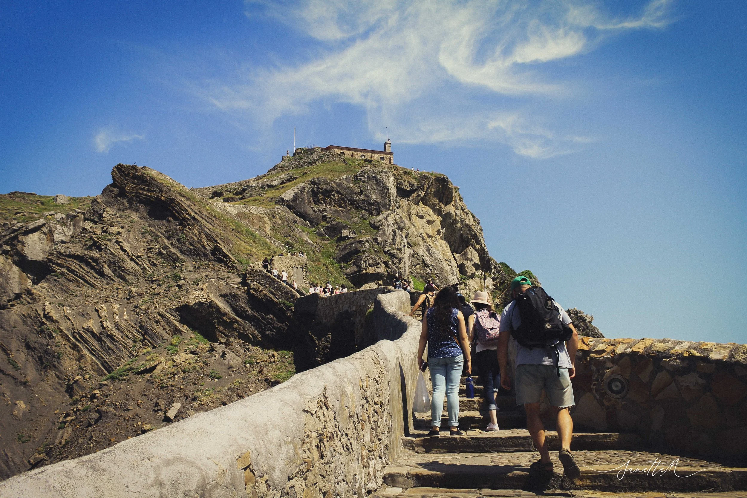 Grupo de personas caminando por un sendero empedrado hacia una cima con un castillo en la cima, en un entorno montañoso con cielo azul y nubes.