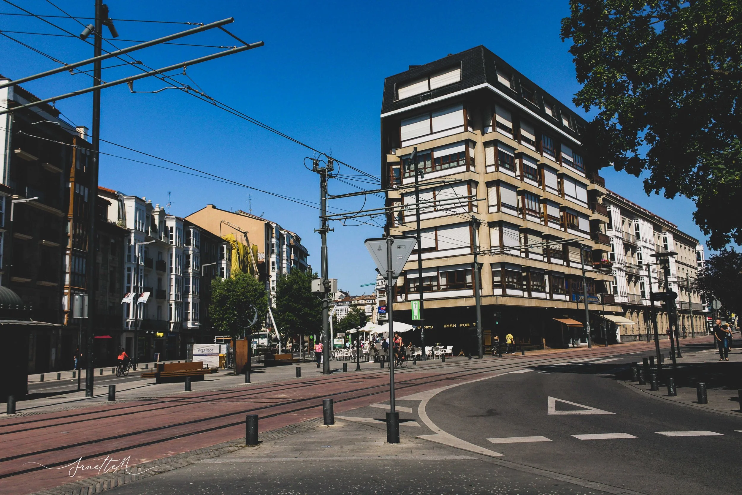 Vista urbana durante el día con edificios de múltiples pisos, postes de electricidad, líneas de tranvía y calles con algunos peatones y ciclistas.