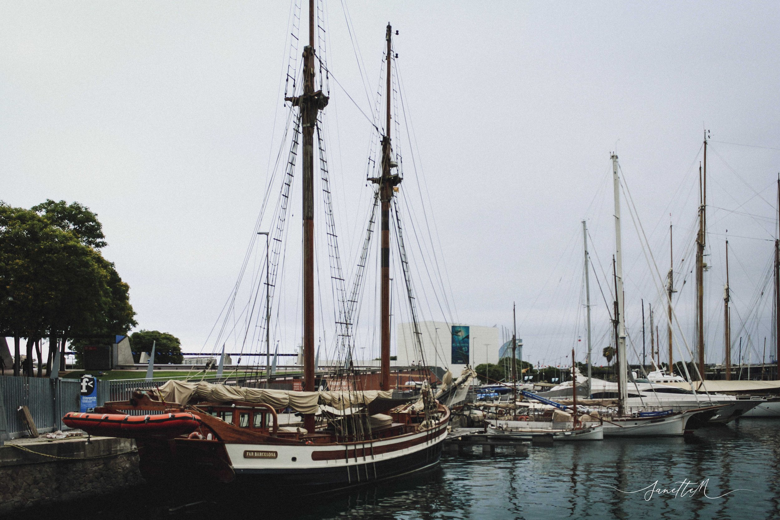 Barcelona - Barcos amarrados en un muelle junto al mar con árboles en el fondo y cielo nublado.
