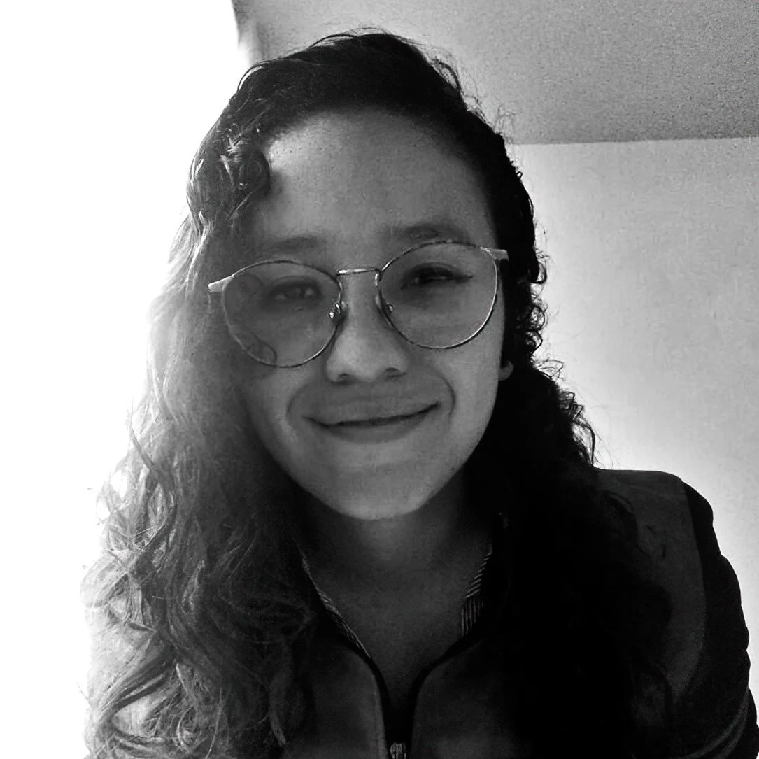 Retrato de una mujer con lentes redondos, cabello rizado y sonrisa en un ambiente interior, en blanco y negro.