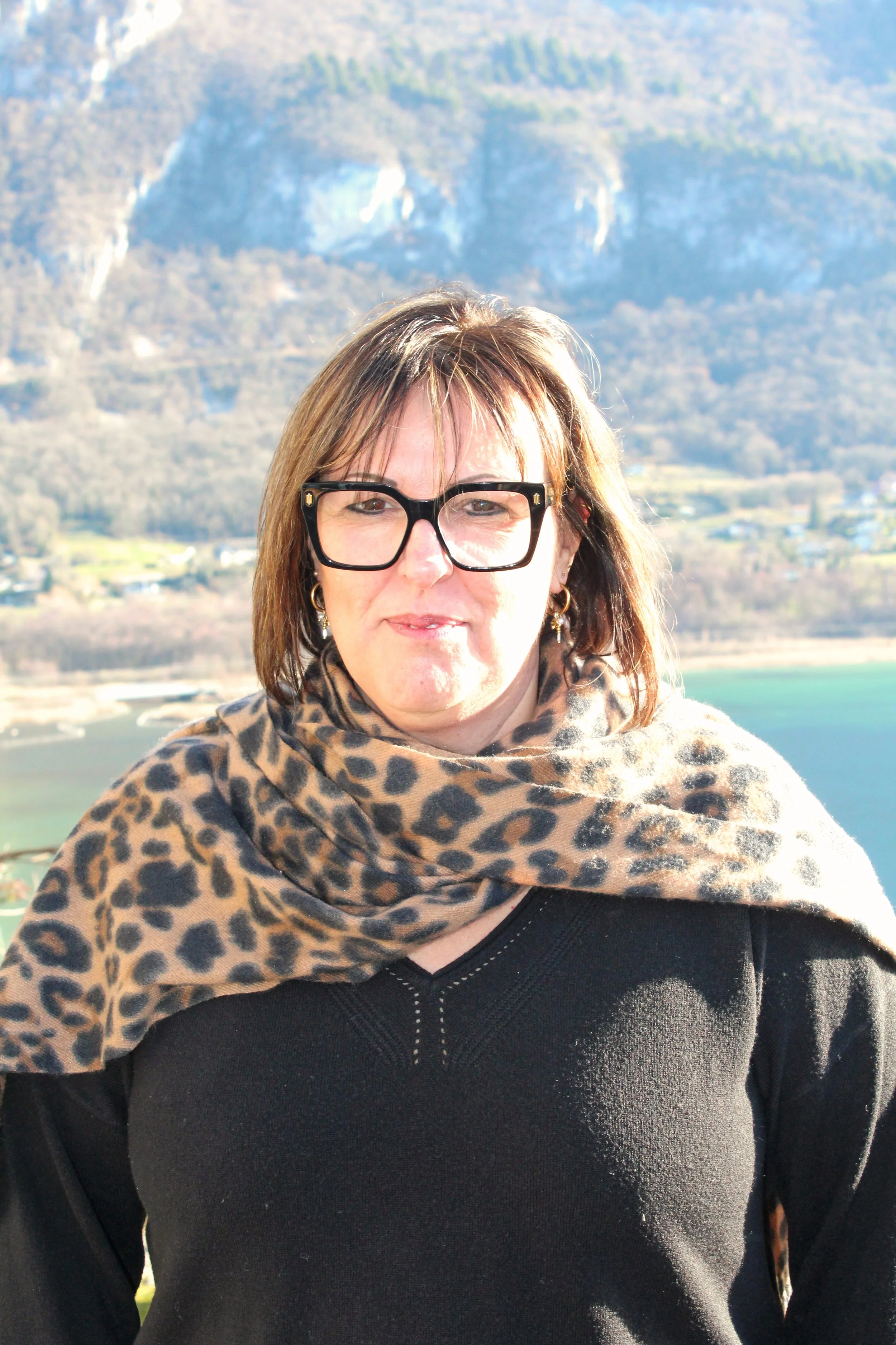 Une femme de Chindrieux portant des lunettes noires et une écharpe à motif léopard, avec un paysage de montagnes, de lac et de campagne en arrière-plan.