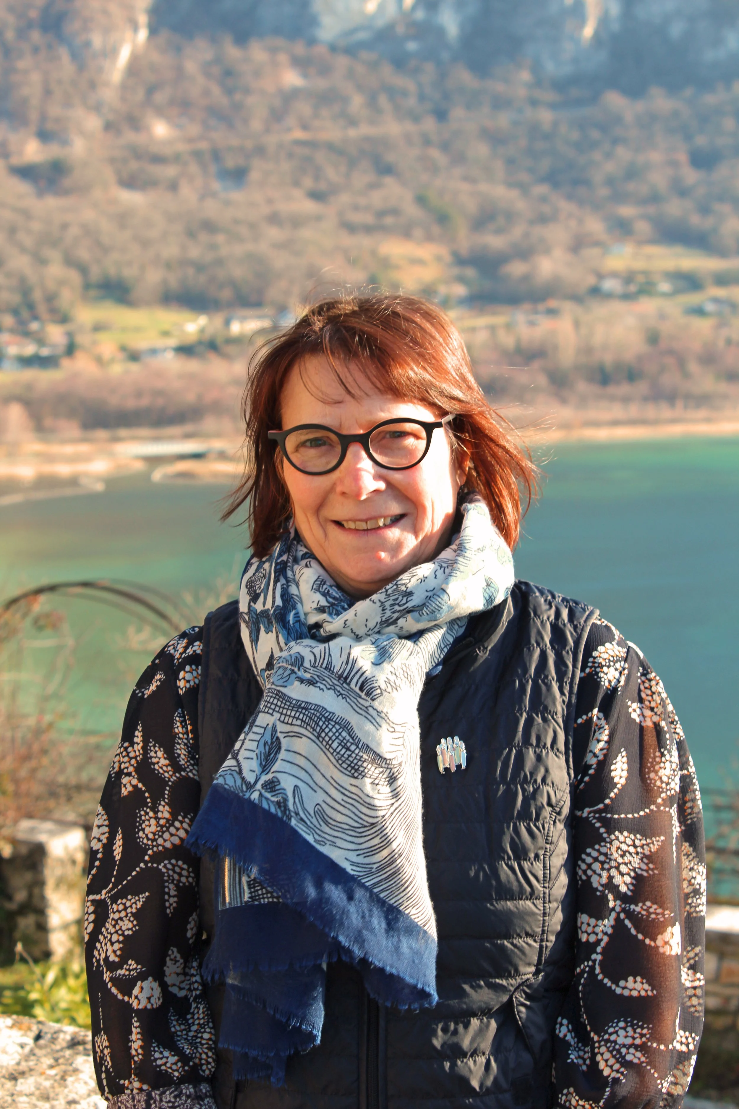 Une femme de Chindrieux souriante portant des lunettes noires, un foulard distinctif et une veste, avec un paysage montagneux et un lac en arrière-plan en plein jour.