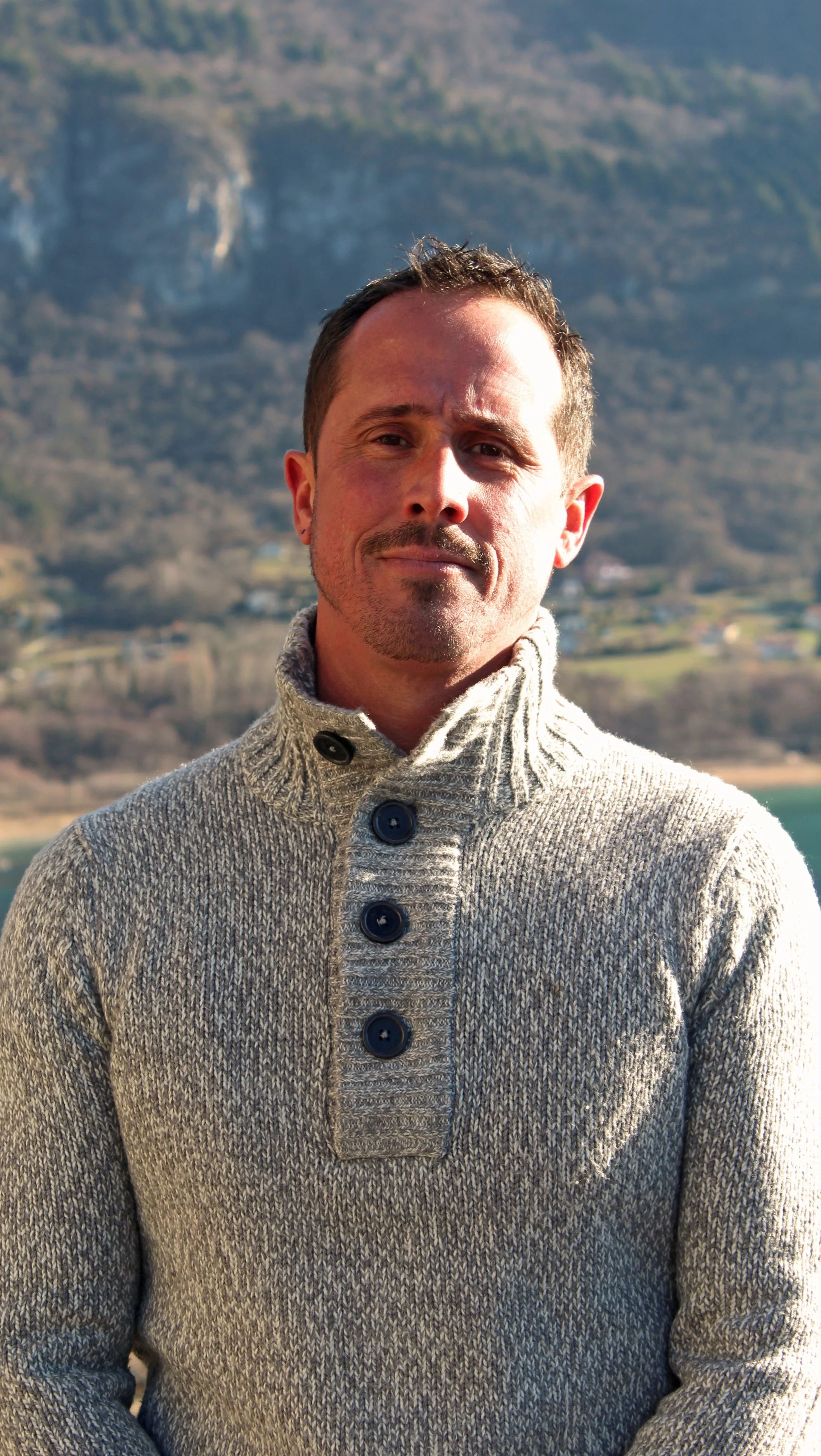 Un homme de Chindrieux portant un pull gris à col roulé avec boutons, debout à l'extérieur avec un paysage montagneux en arrière-plan.