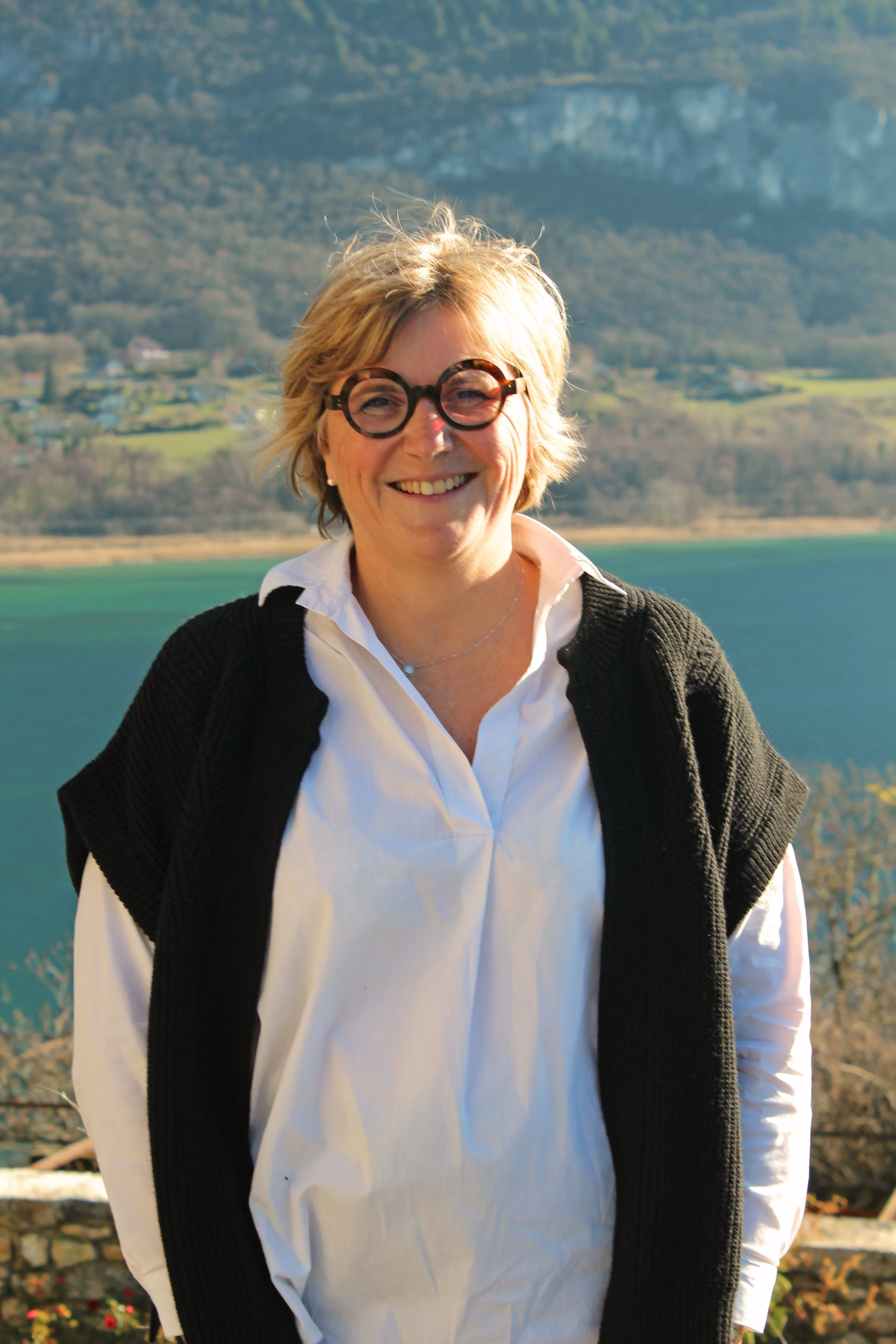 Femme de Chindrieux souriante portant des lunettes rondes, un chemisier blanc et un gilet noir, avec un paysage de lac et de montagnes en arrière-plan.