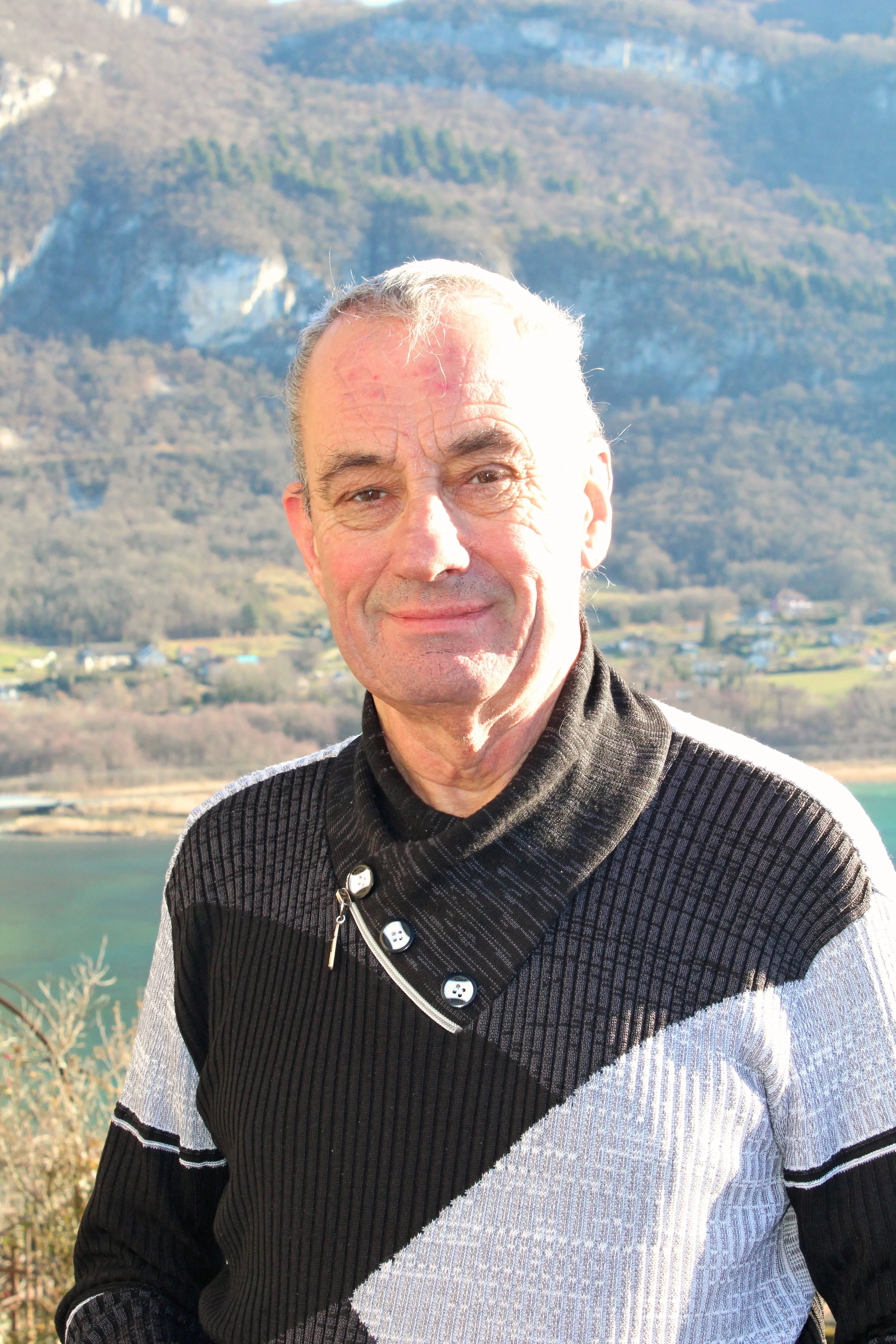 Un homme de Chindrieux âgé souriant devant un paysage de montagnes et un lac, avec des terrains verdoyants en arrière-plan.