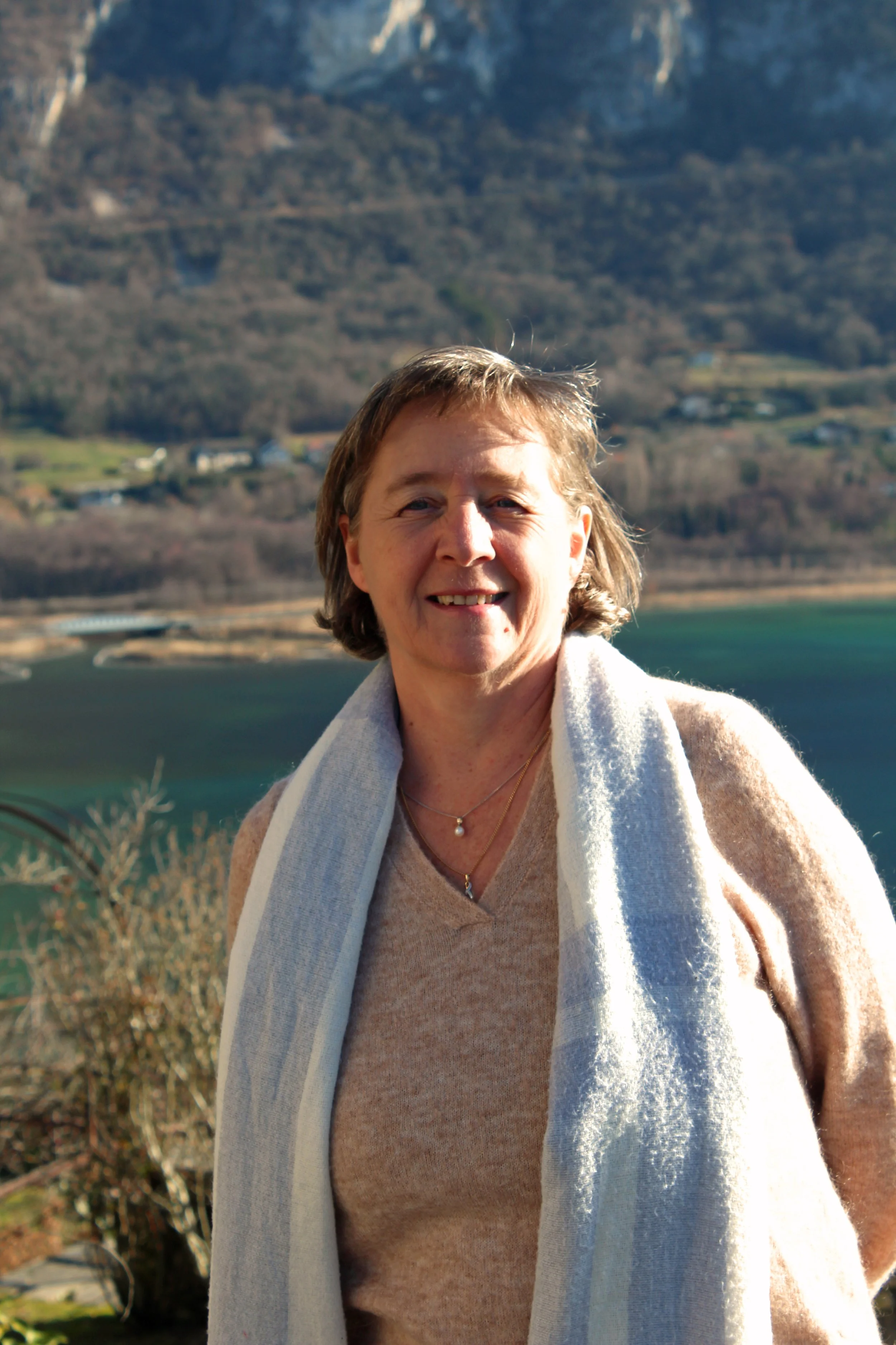 Mari-Claire BARBIER debout en plein air avec un lac et des montagnes en arrière-plan, ensoleillé.