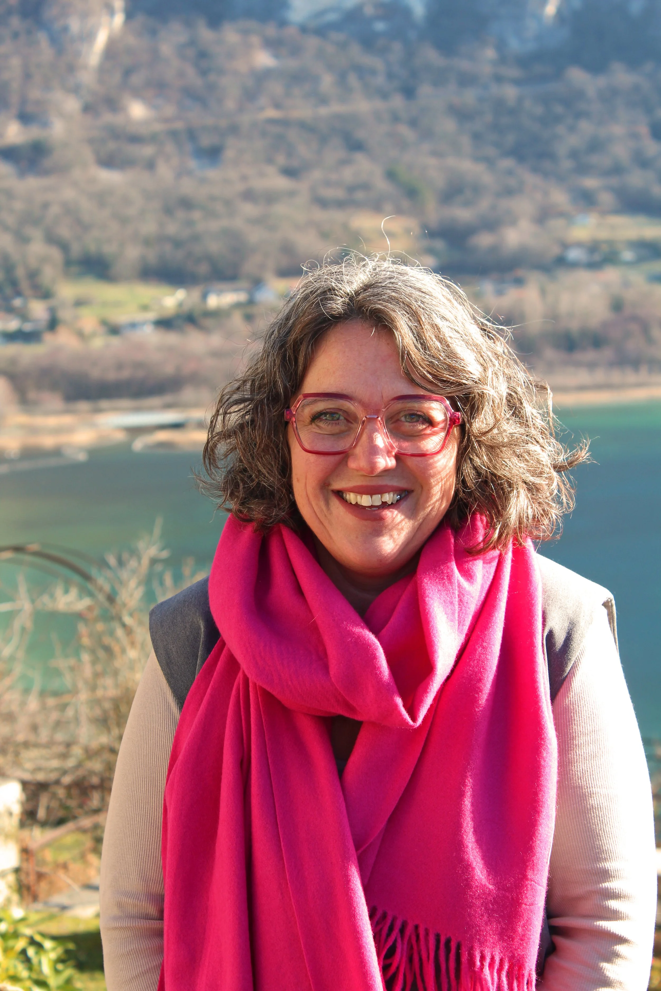 Femme de Chindrieux souriante portant des lunettes roses et une écharpe rose, avec un paysage naturel en arrière-plan.