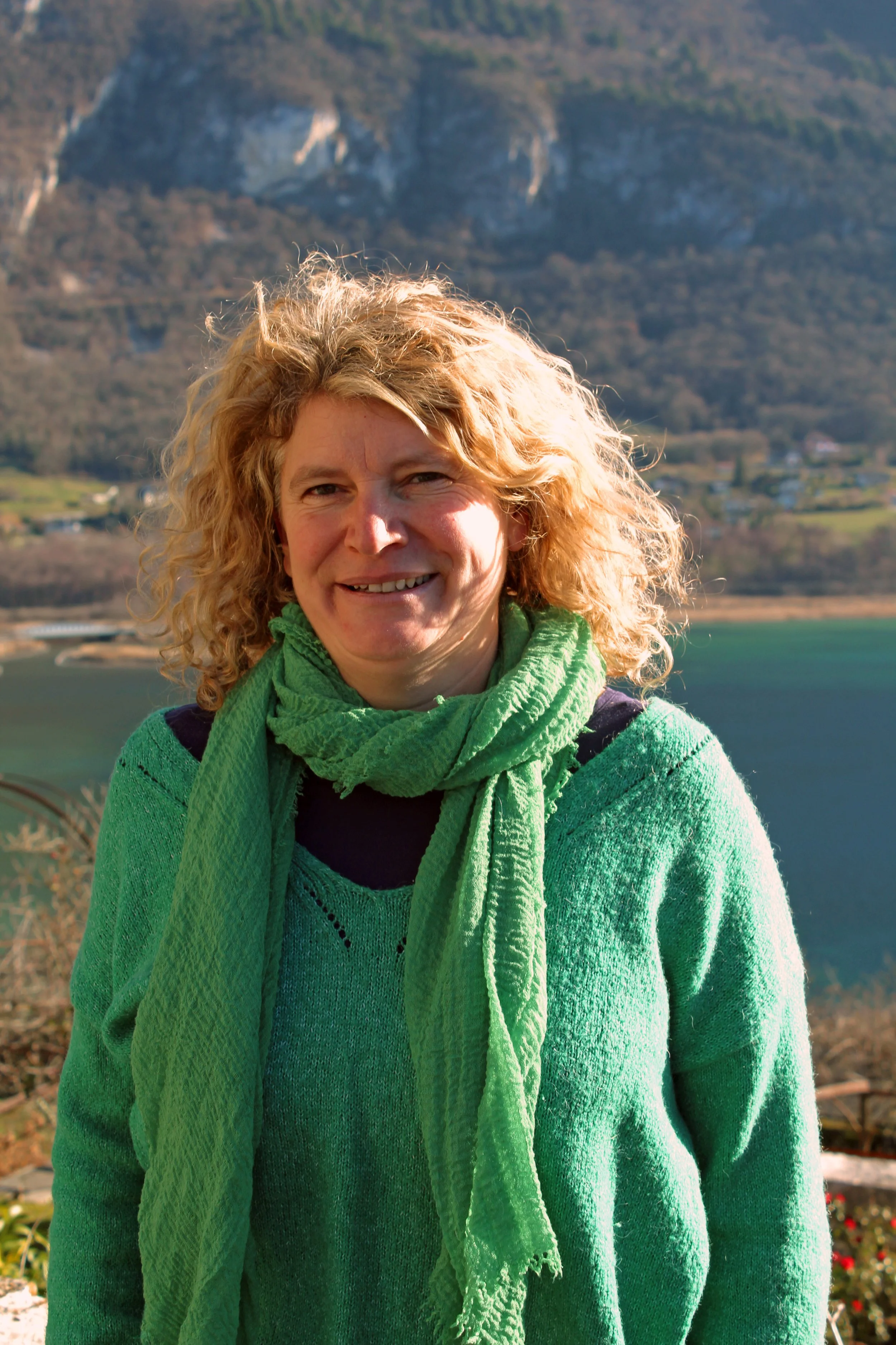 Une femme de Chindrieux souriante avec des cheveux bouclés, portant un pull vert et une écharpe verte, en plein air avec un lac et des montagnes en arrière-plan.