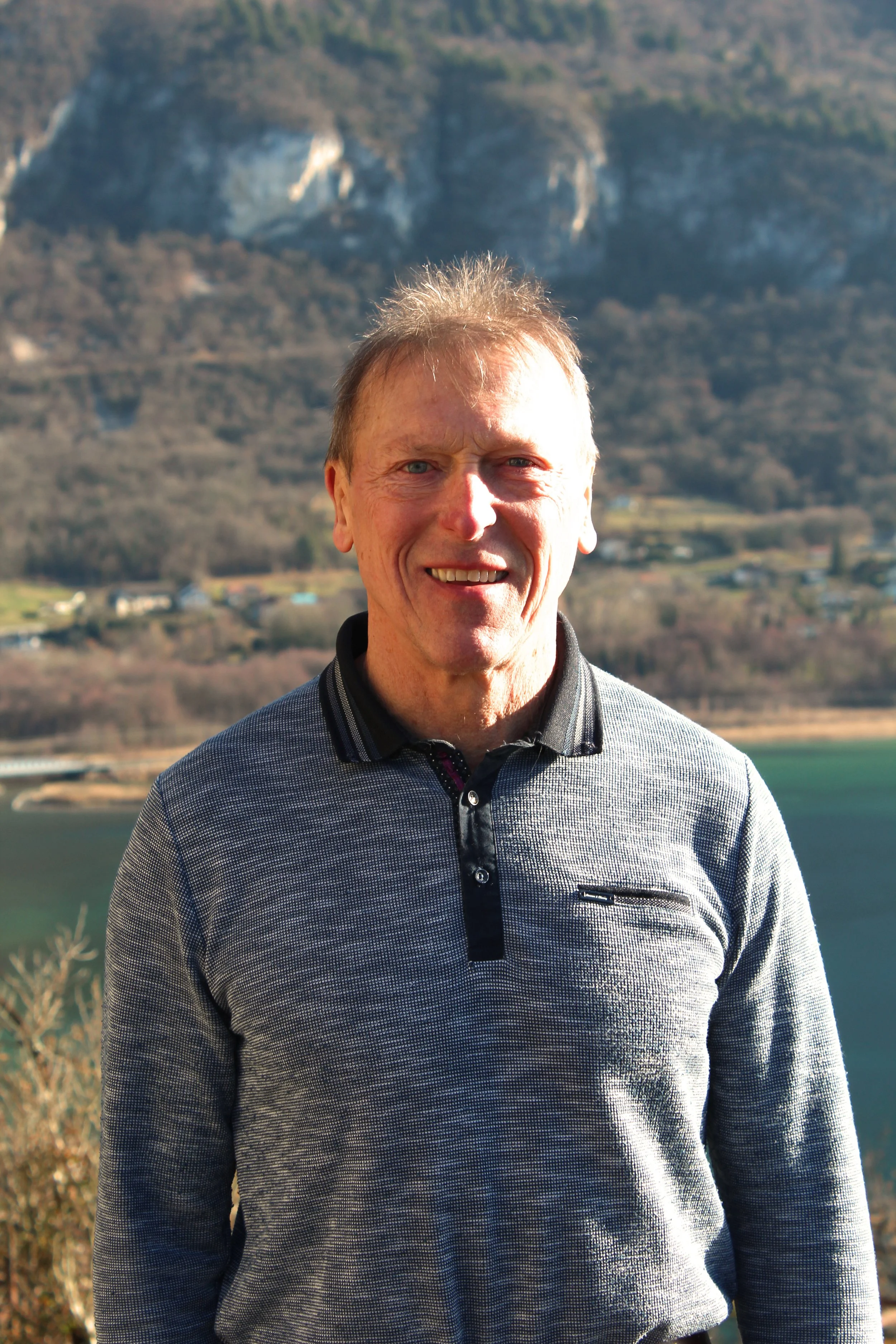 Un homme de Chindrieux souriant en pull gris, avec un paysage de montagnes et d'eau en arrière-plan.