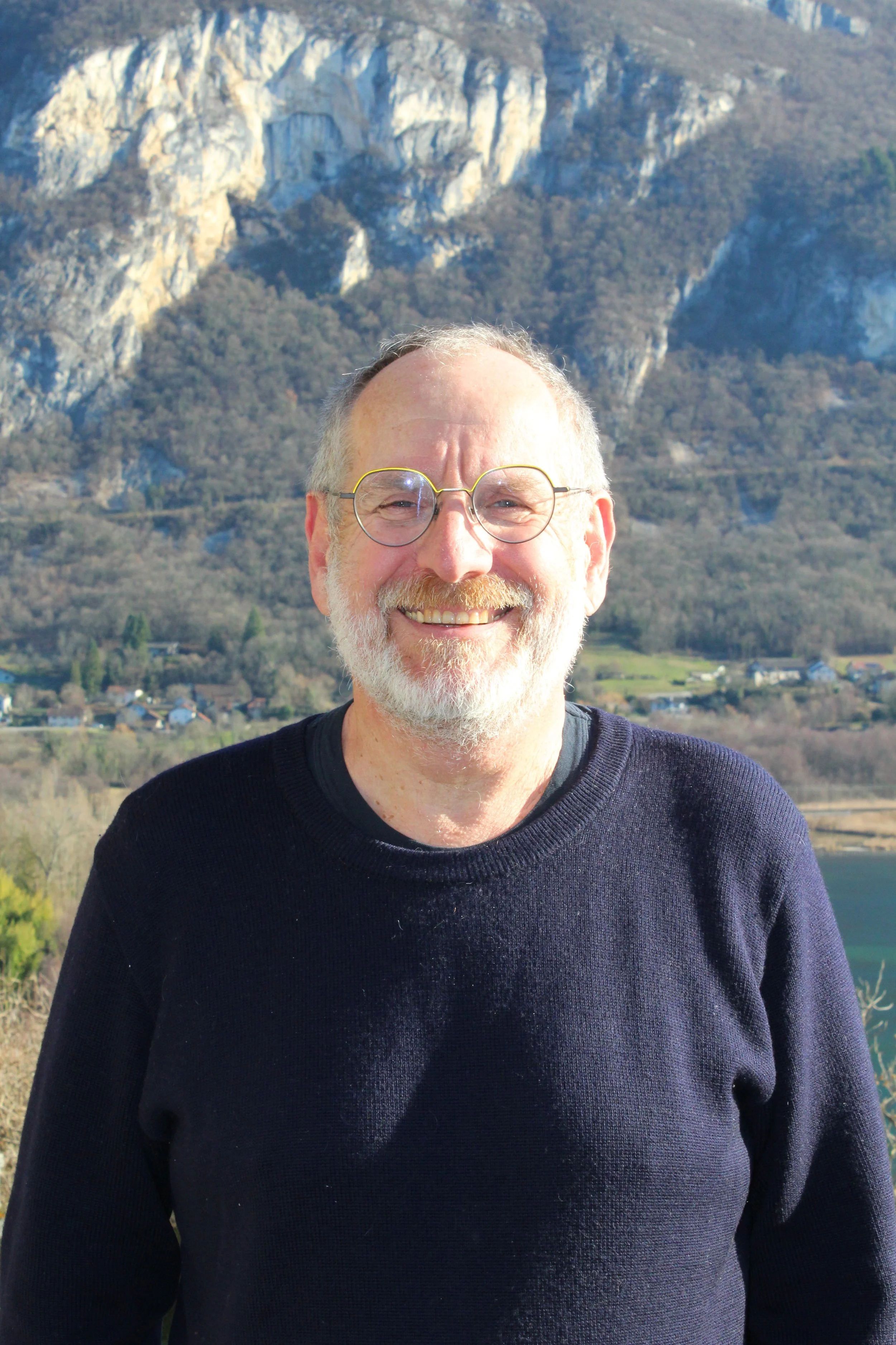 Homme de Chindrieux souriant portant des lunettes, vêtu d'un pull noir, avec un paysage montagneux en arrière-plan.