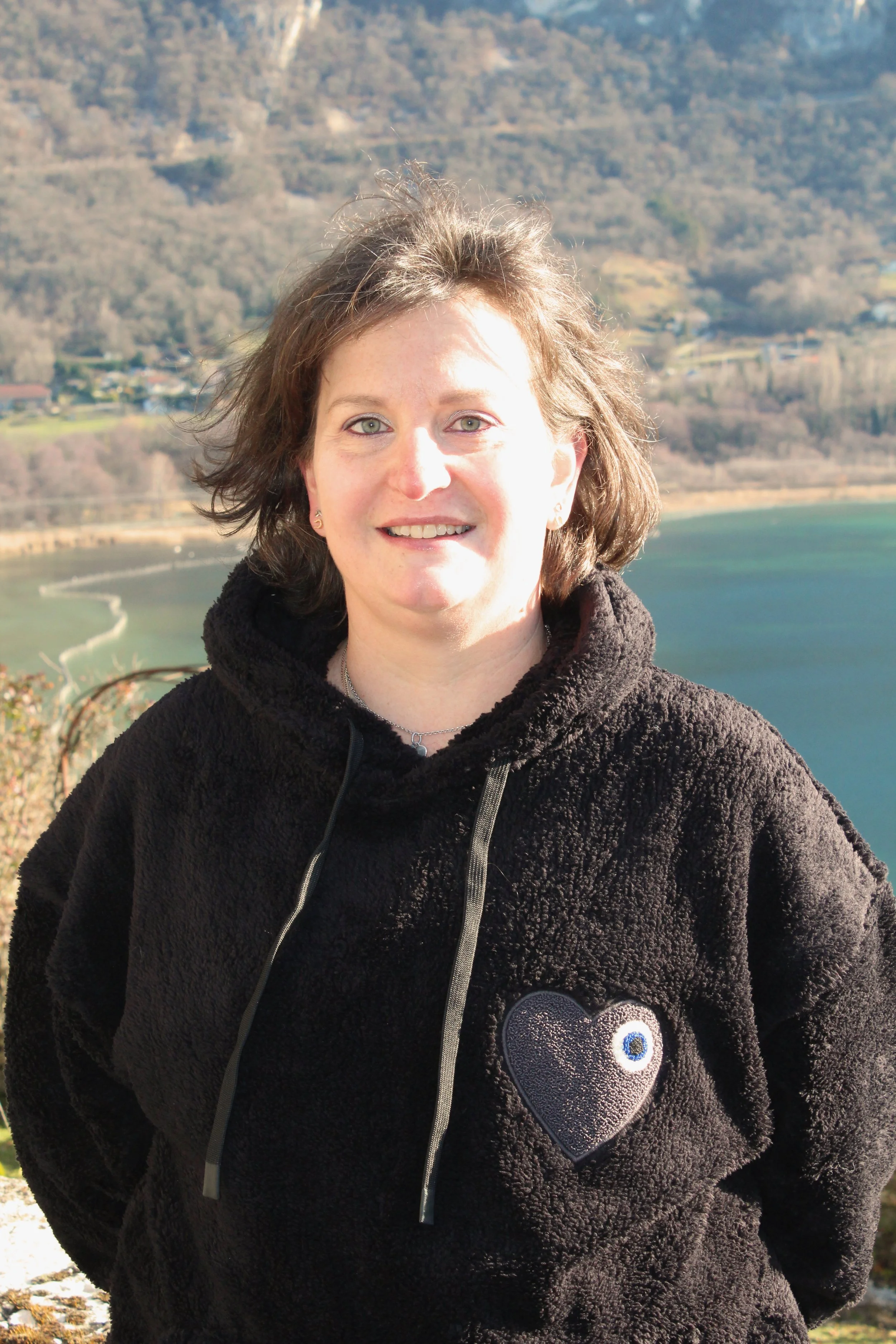 Une femme de Chindrieux souriante avec des cheveux bruns courts, portant un pull noir avec un cœur brodé contenant un œil, en extérieur avec un lac et des montagnes en arrière-plan.