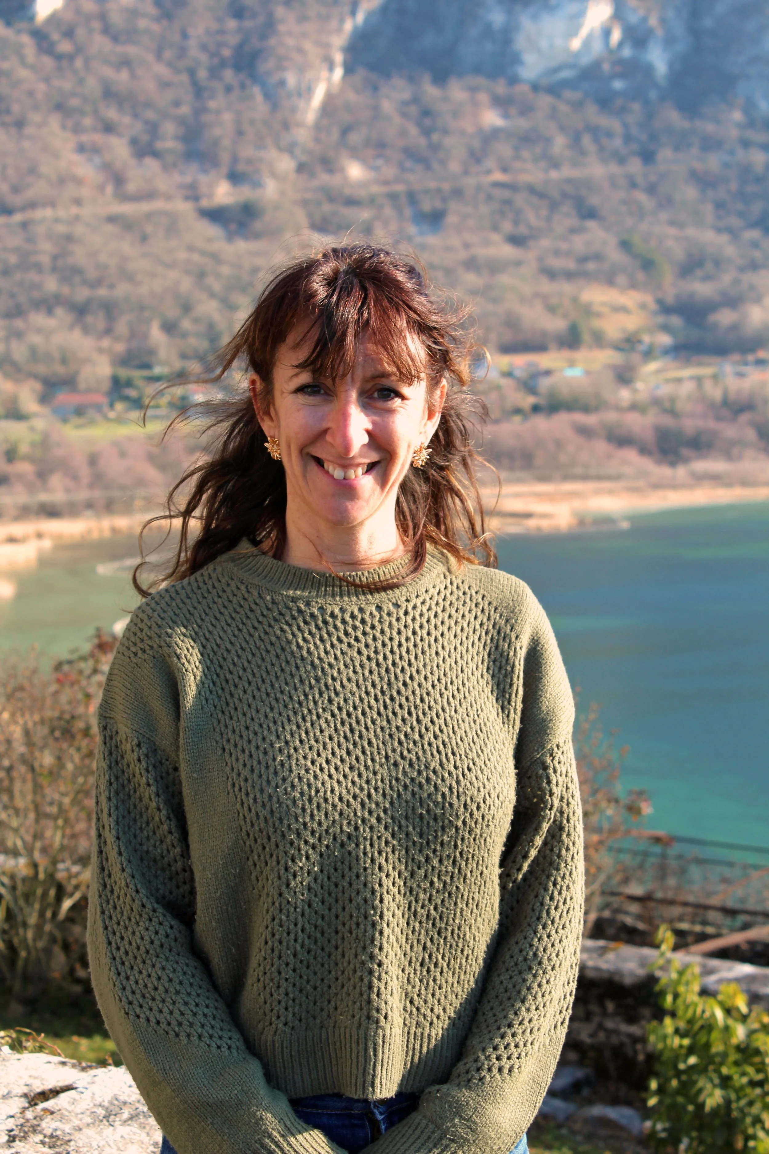 Une femme de Chindrieux souriante avec des cheveux bruns ondulés, portant un pull vert olive, devant un panorama de montagnes et un lac en arrière-plan.