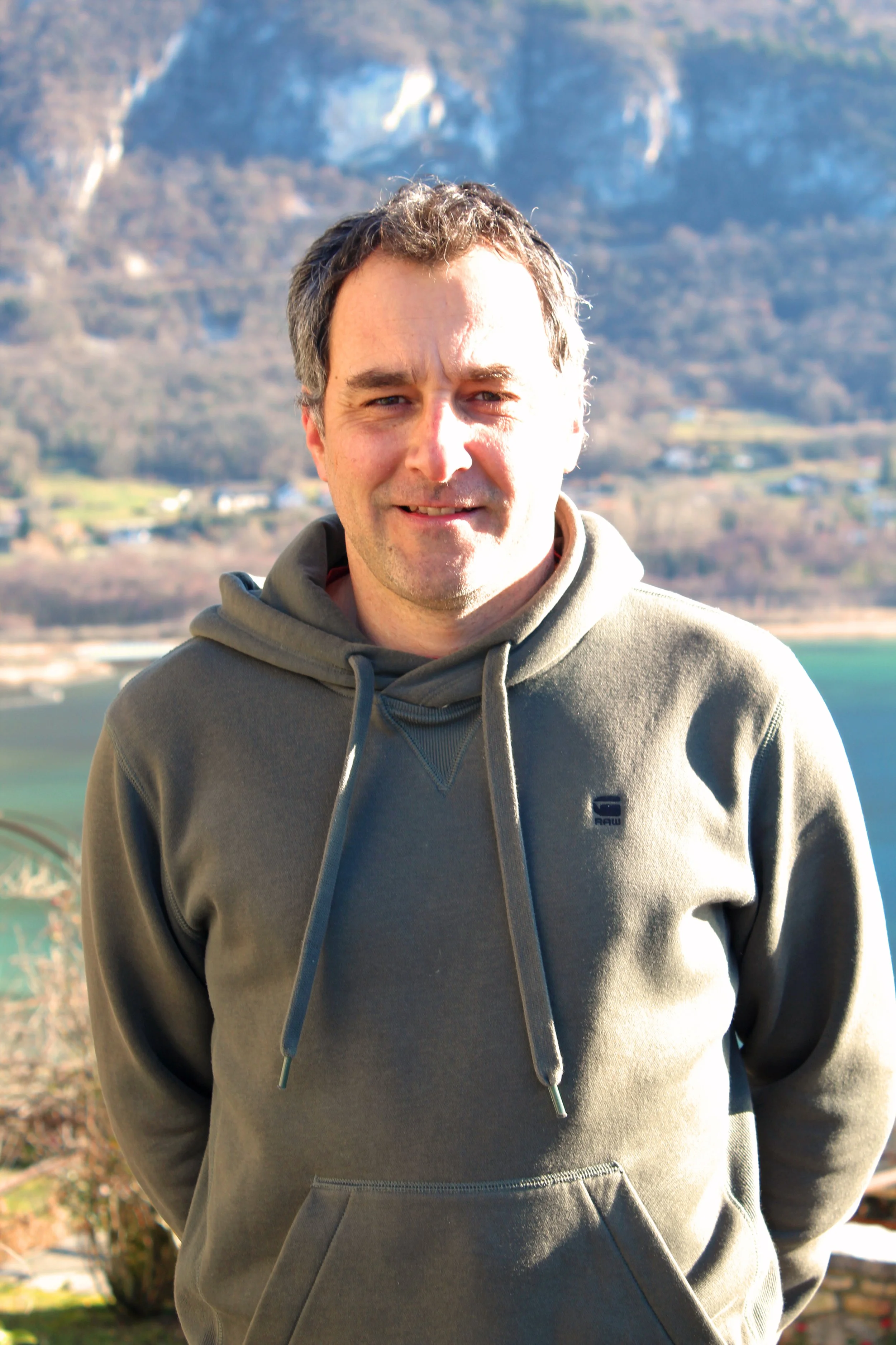 Un homme de Chindrieux souriant portant un sweat à capuche gris, avec un paysage de montagnes et un lac en arrière-plan.