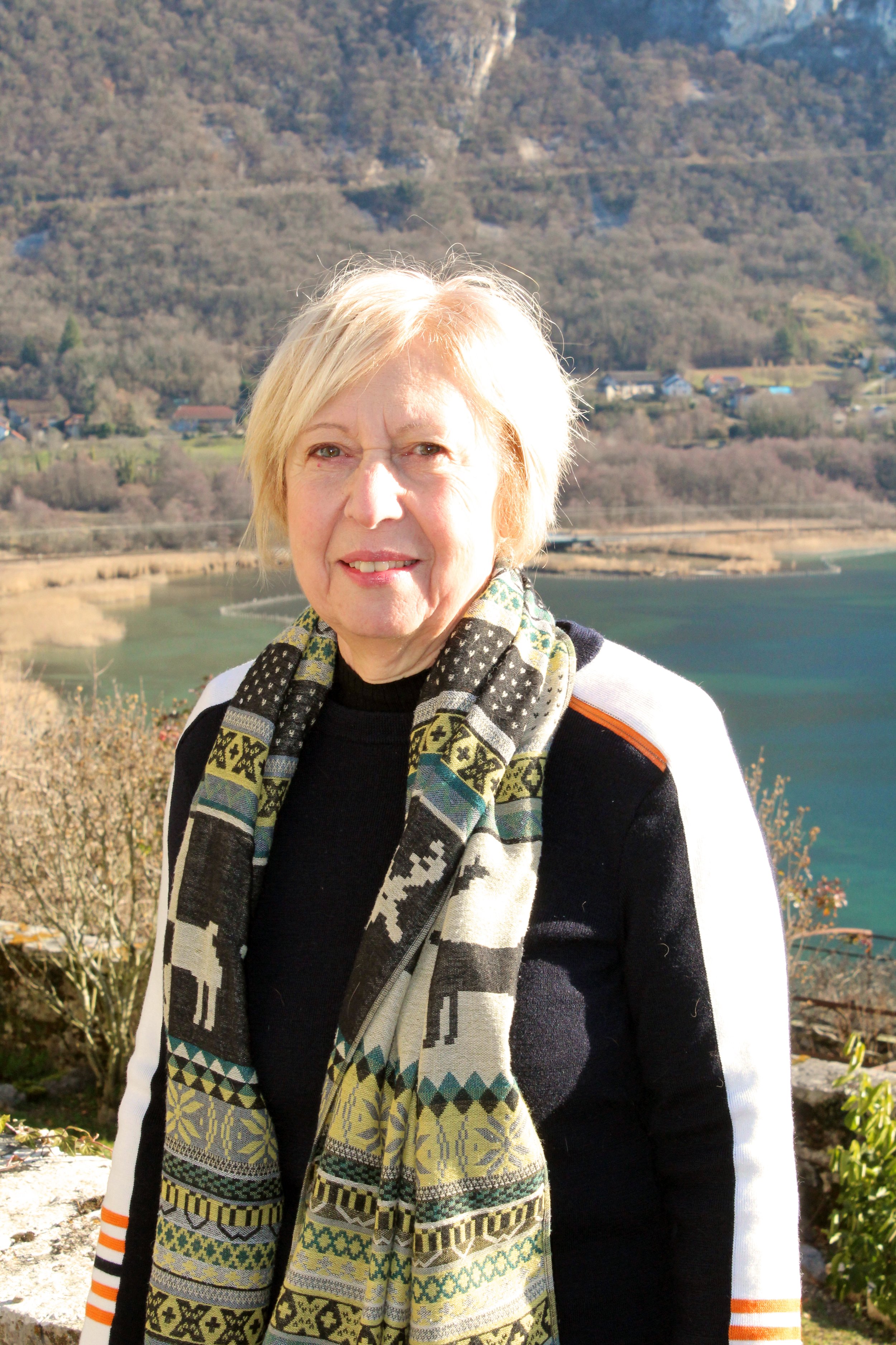 Une femme de Chindrieux avec des cheveux blonds courts, portant un foulard coloré, devant un paysage de montagnes, lac et végétation en arrière-plan.