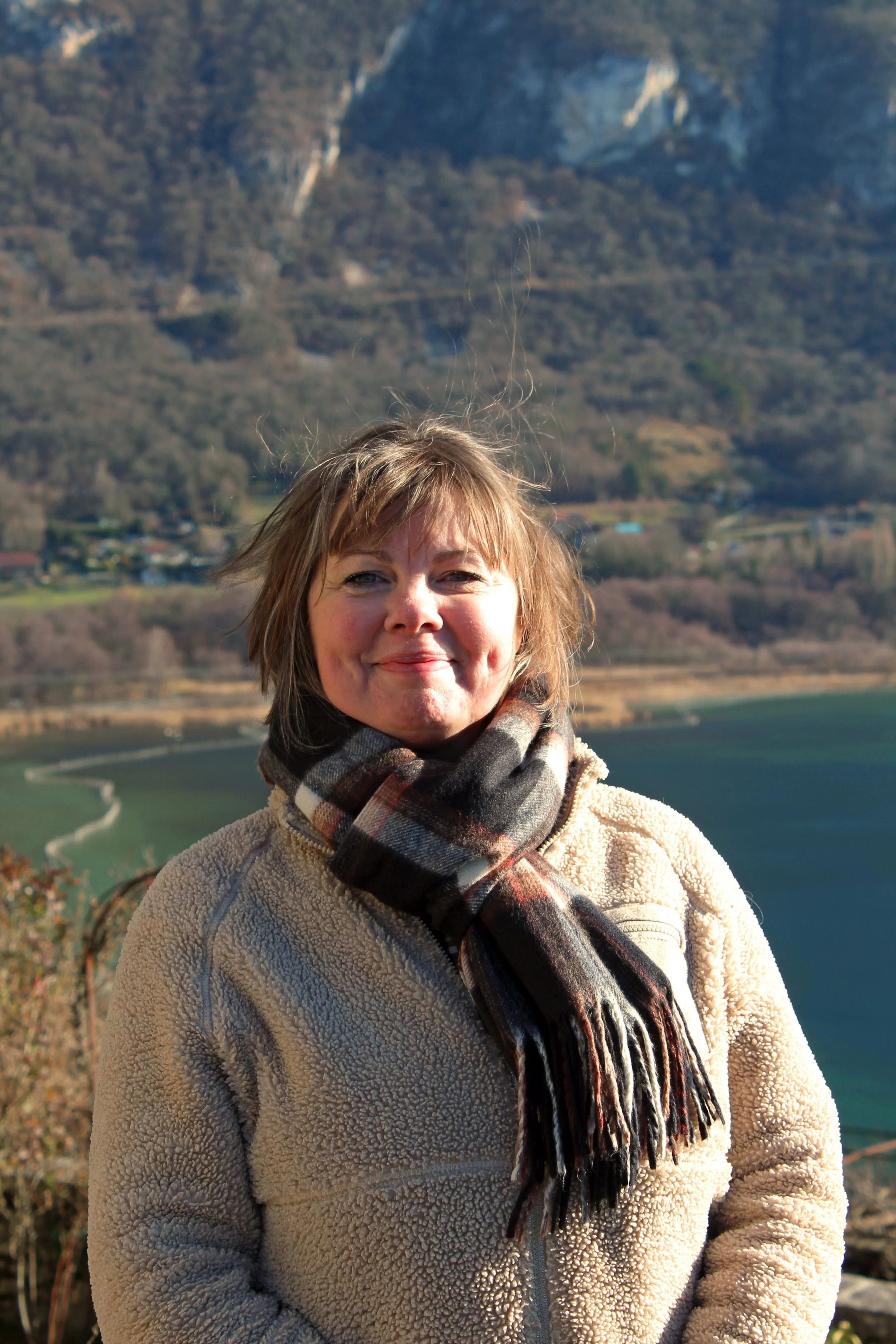 Une femme Chindrieux souriante, portant un manteau beige en peluche et une écharpe à carreaux, se tient devant un paysage naturel avec un lac, des collines et des montagnes en arrière-plan.